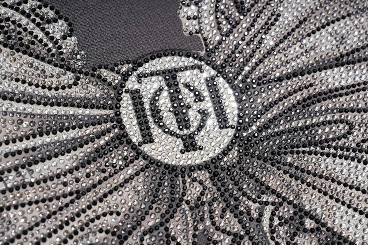 Close-up of a black and silver embellished garment featuring a central circular monogram logo with the letters 'T', 'C', and 'A' surrounded by dense rhinestone or beadwork on a dark gray background. TCH Rhinestone Bow Graphic Fitted Long Sleeve Top