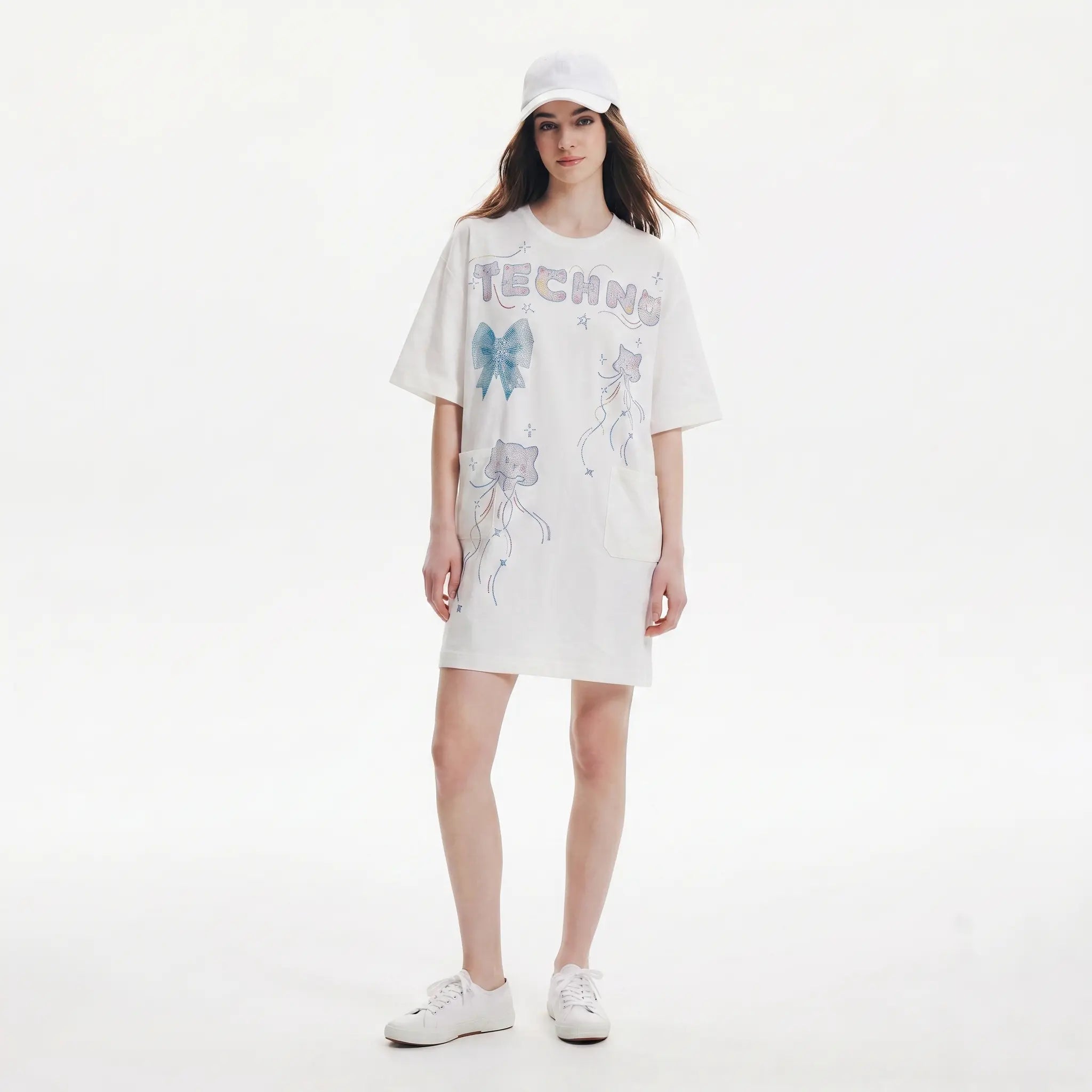 A model wearing an oversized white graphic T-shirt dress with 'TECHNO' and cat illustrations, styled with a white cap and white sneakers against a plain white background. TCH Rhinestone Bow Graphic Oversized T-Shirt Dress