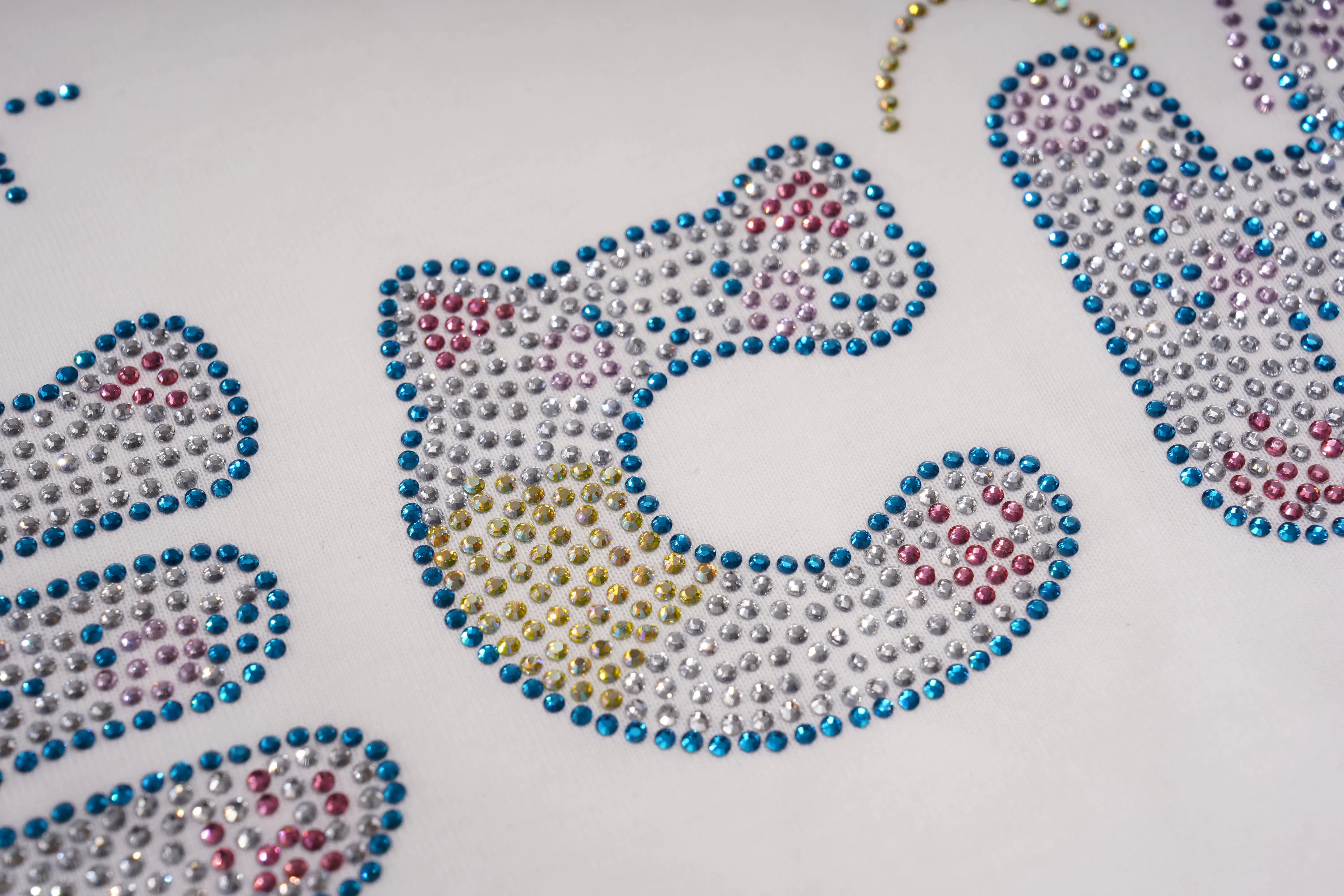 A close-up of a white surface with several decorative letter or shape appliqués covered in small rhinestones. The central piece is a curved shape outlined in blue and filled with silver, pink, and gold rhinestones, with other matching rhinestone decorations partially visible around it. TCH Rhinestone Bow Graphic Oversized T-Shirt Dress