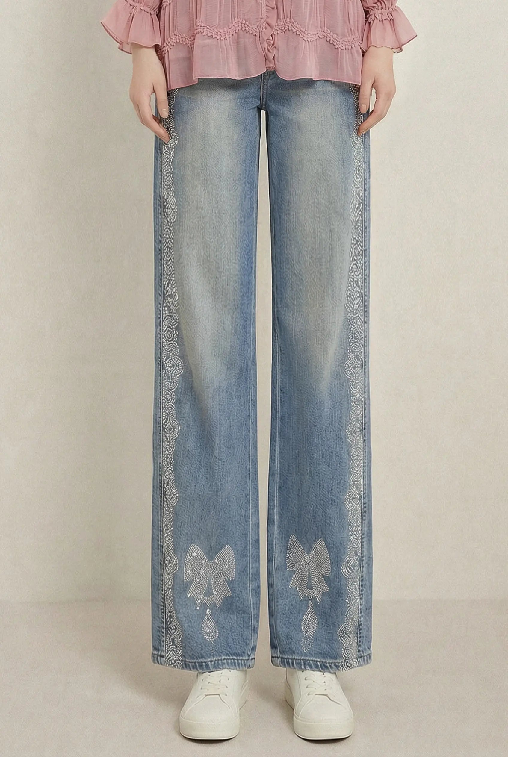 A person wearing light blue wide-leg jeans with white lace trim down the sides and white sneakers, paired with a pink ruffled blouse, against a plain beige studio background. TCH Rhinestone Bow Lace Side Panel Wide Leg Jeans