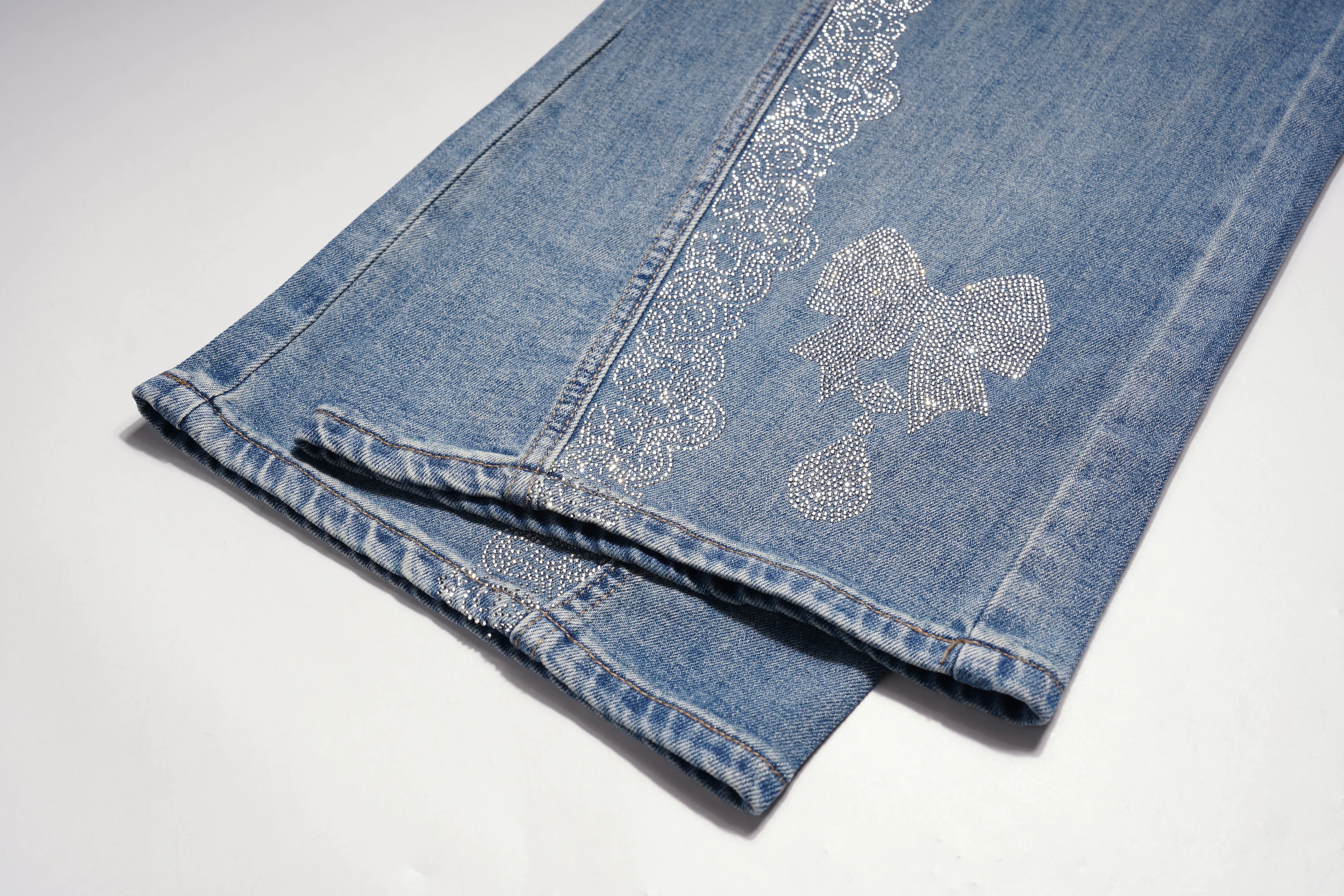 Light blue denim jeans folded on a white background, featuring intricate silver rhinestone embroidery along the leg and a matching embroidered patch near the knee. TCH Rhinestone Bow Lace Side Panel Wide Leg Jeans