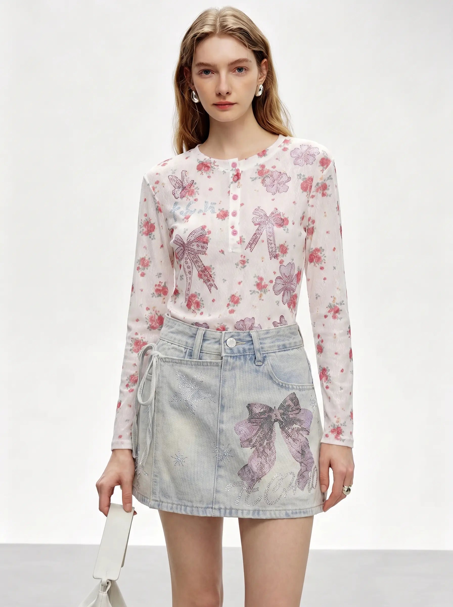 A model wearing a sheer long-sleeve button-up top with a pink and purple bow and floral print, paired with a light-wash denim mini skirt featuring a large bow graphic, against a plain light gray studio background. TCH Rhinestone Bow Light Wash Denim Shorts
