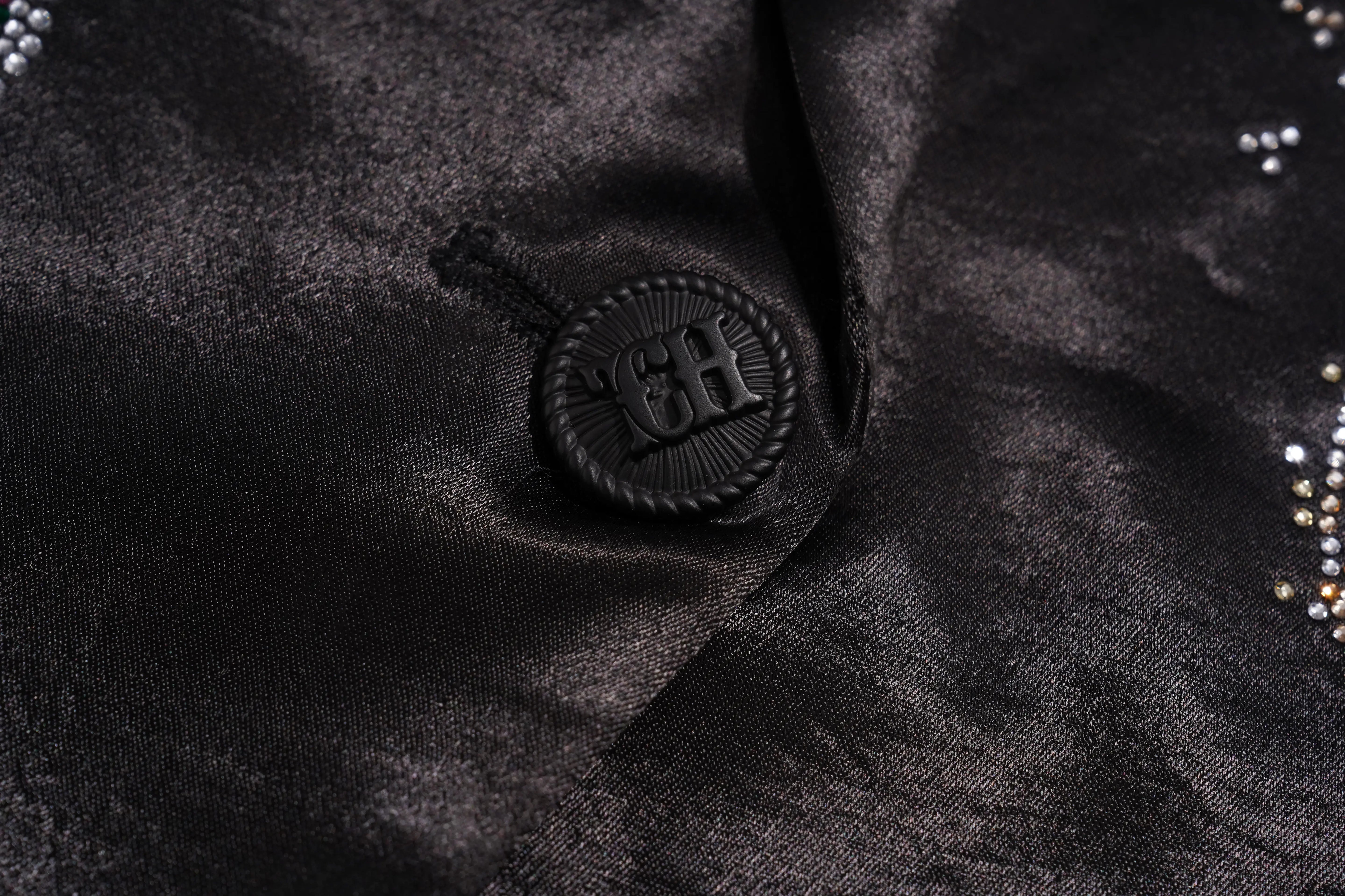 Close-up of a black satin or silk-like garment surface with a round black decorative button featuring an embossed 'CH' monogram, plus small rhinestone embellishments along the edge. TCH Rhinestone Bow Satin Blazer