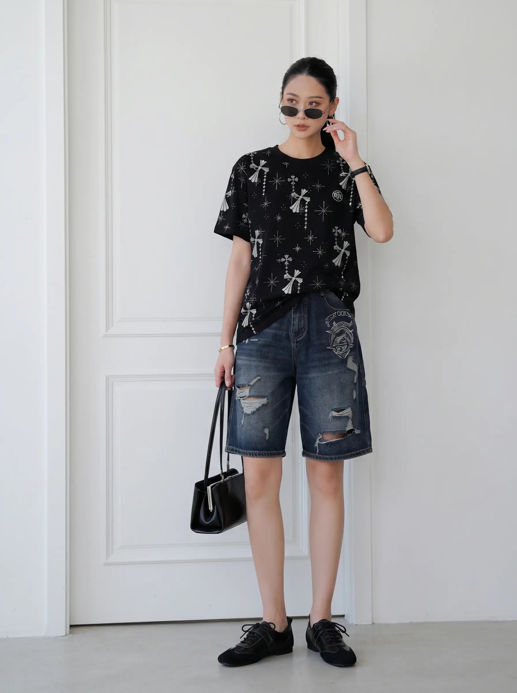 A woman stands indoors against a white paneled wall wearing a black graphic T-shirt with white cross motifs, distressed blue denim shorts, black shoes, and holding a black handbag. TCH Rhinestone Bow Star Pattern Black T-Shirt