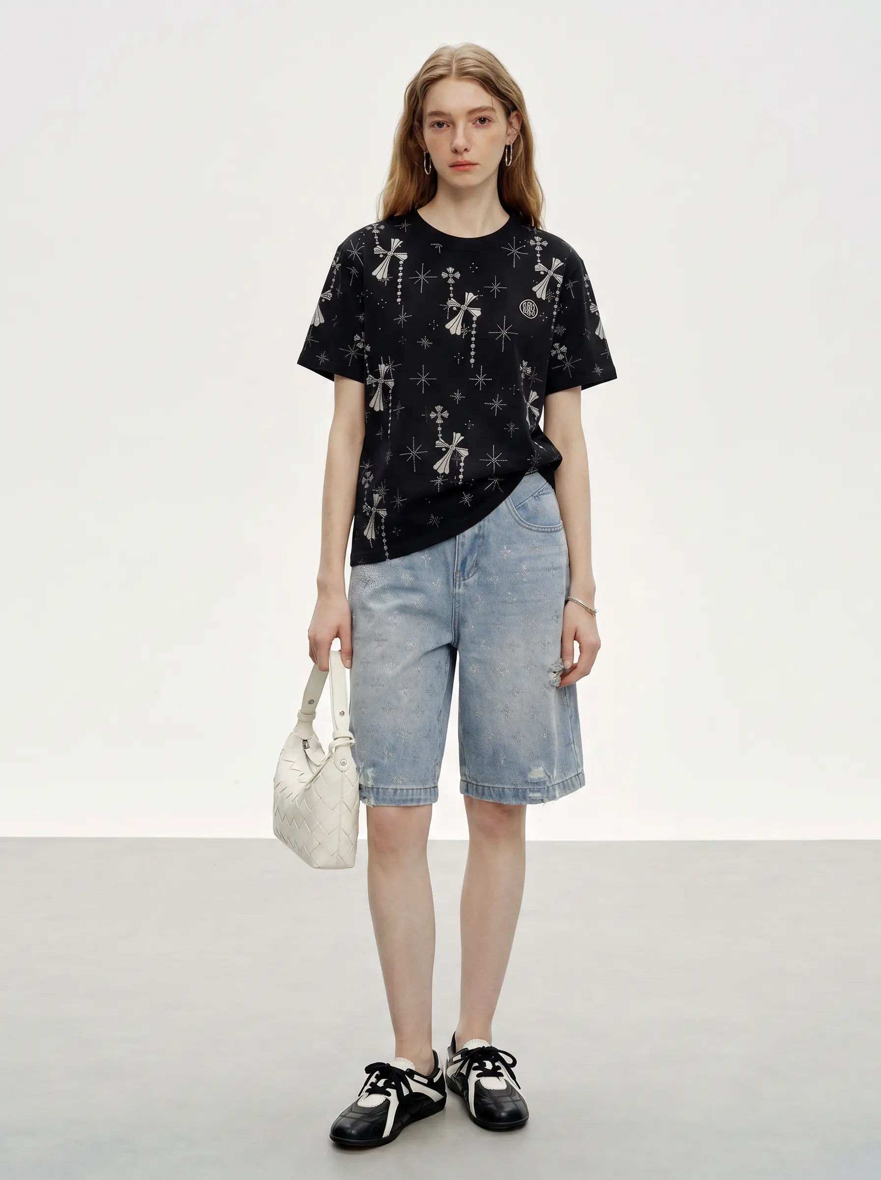 A model stands against a light gray studio background wearing a black short-sleeve t-shirt with a white cross pattern, light-wash denim shorts, black sneakers, and holding a white handbag. TCH Rhinestone Bow Star Pattern Black T-Shirt