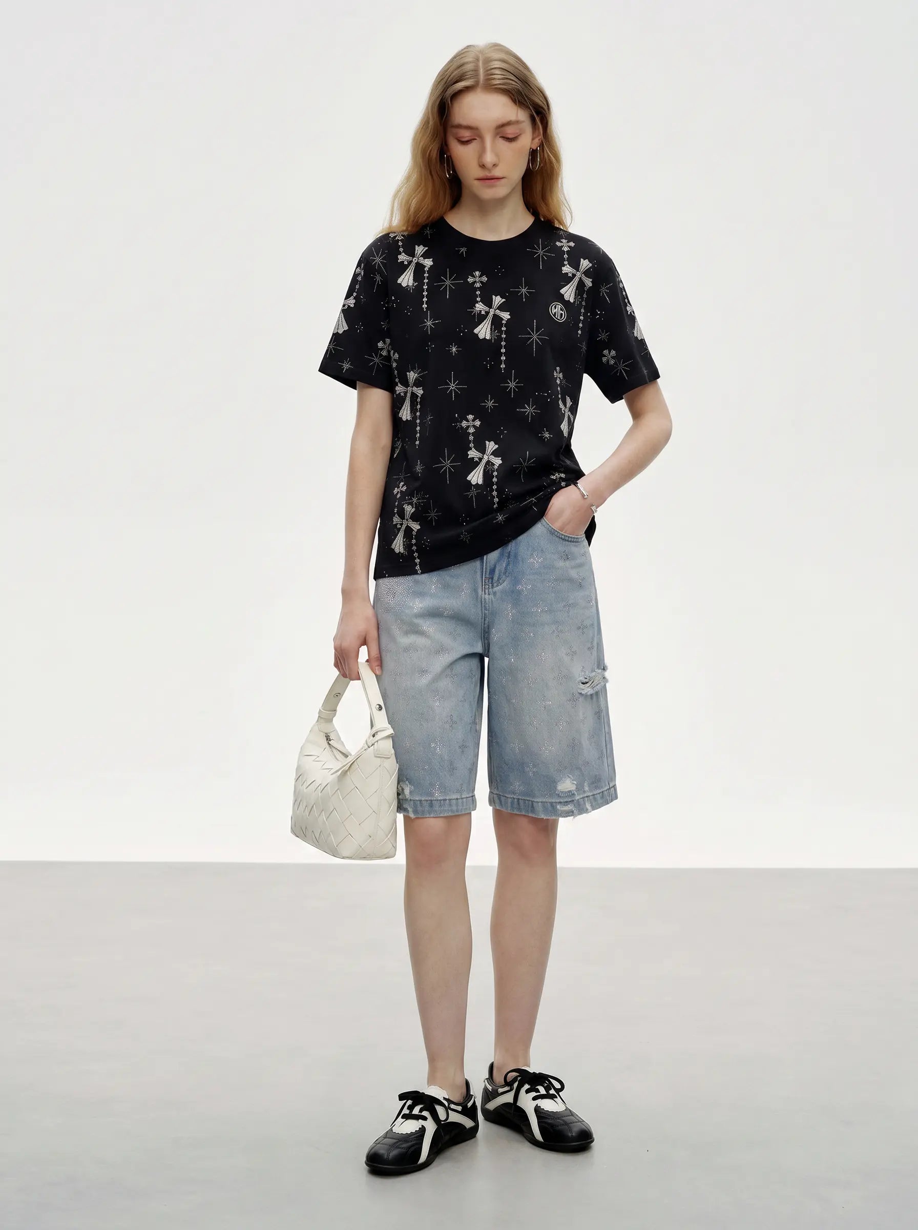 A model stands in a studio wearing a black short-sleeve t-shirt with a white celestial print, light-wash denim shorts, black and white sneakers, and holding a textured cream handbag. TCH Rhinestone Bow Star Pattern Black T-Shirt