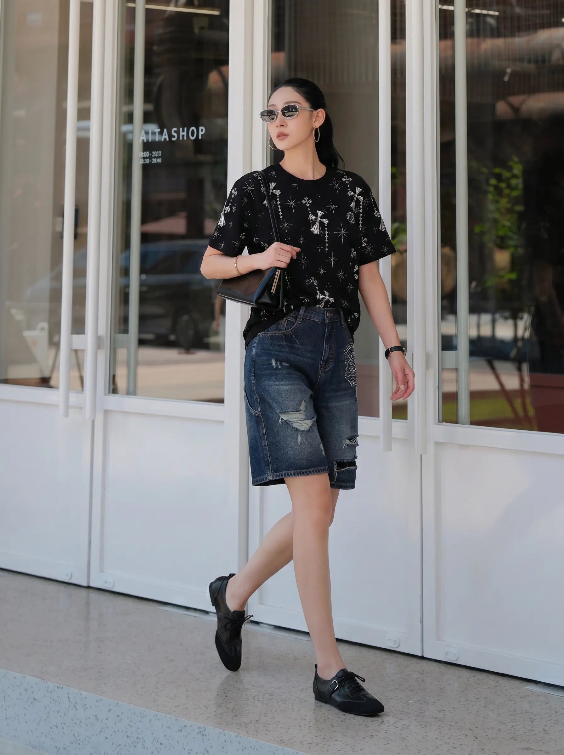 A woman walking outdoors in front of a glass storefront, wearing a black short-sleeve t-shirt with a white graphic print, distressed blue denim shorts, and black loafers. TCH Rhinestone Bow Star Pattern Black T-Shirt