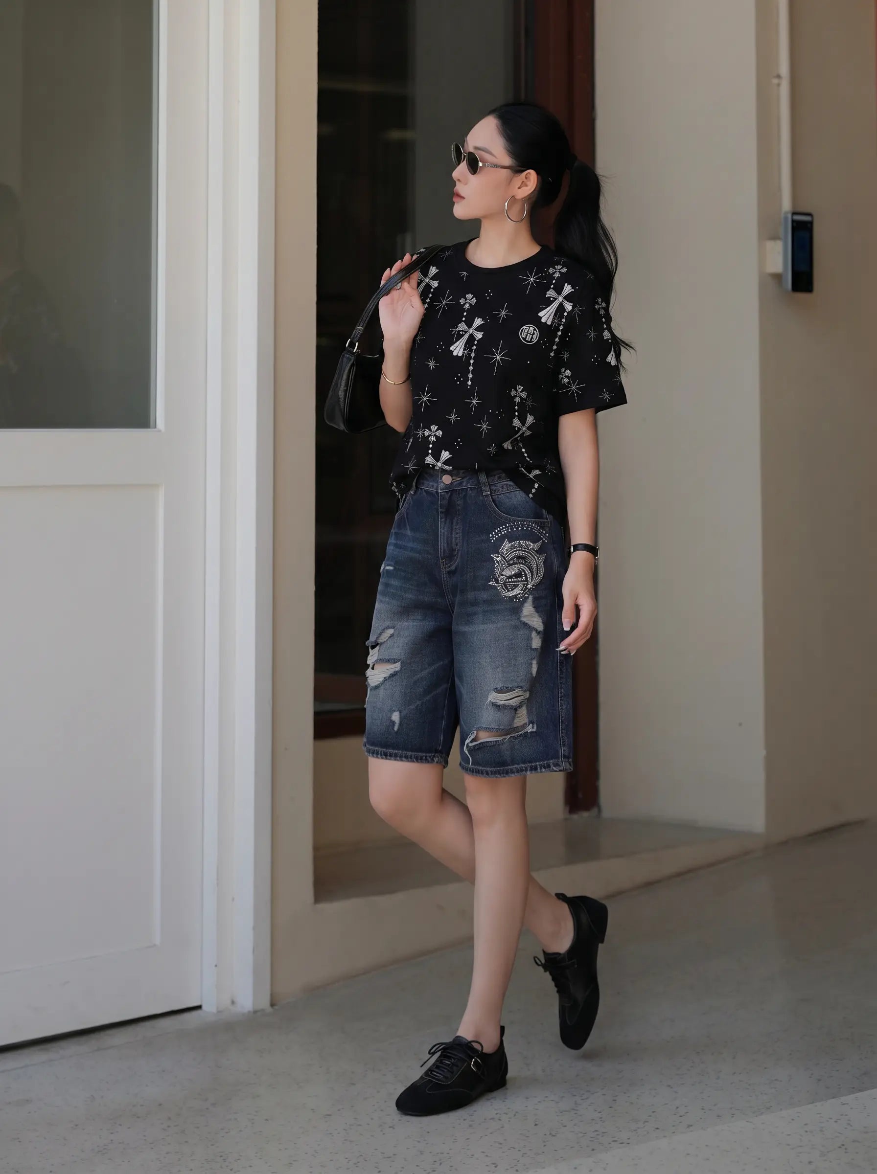 A woman wearing a black short-sleeve t-shirt with white celestial prints and distressed blue denim shorts, walking in a modern indoor setting. TCH Rhinestone Bow Star Pattern Black T-Shirt