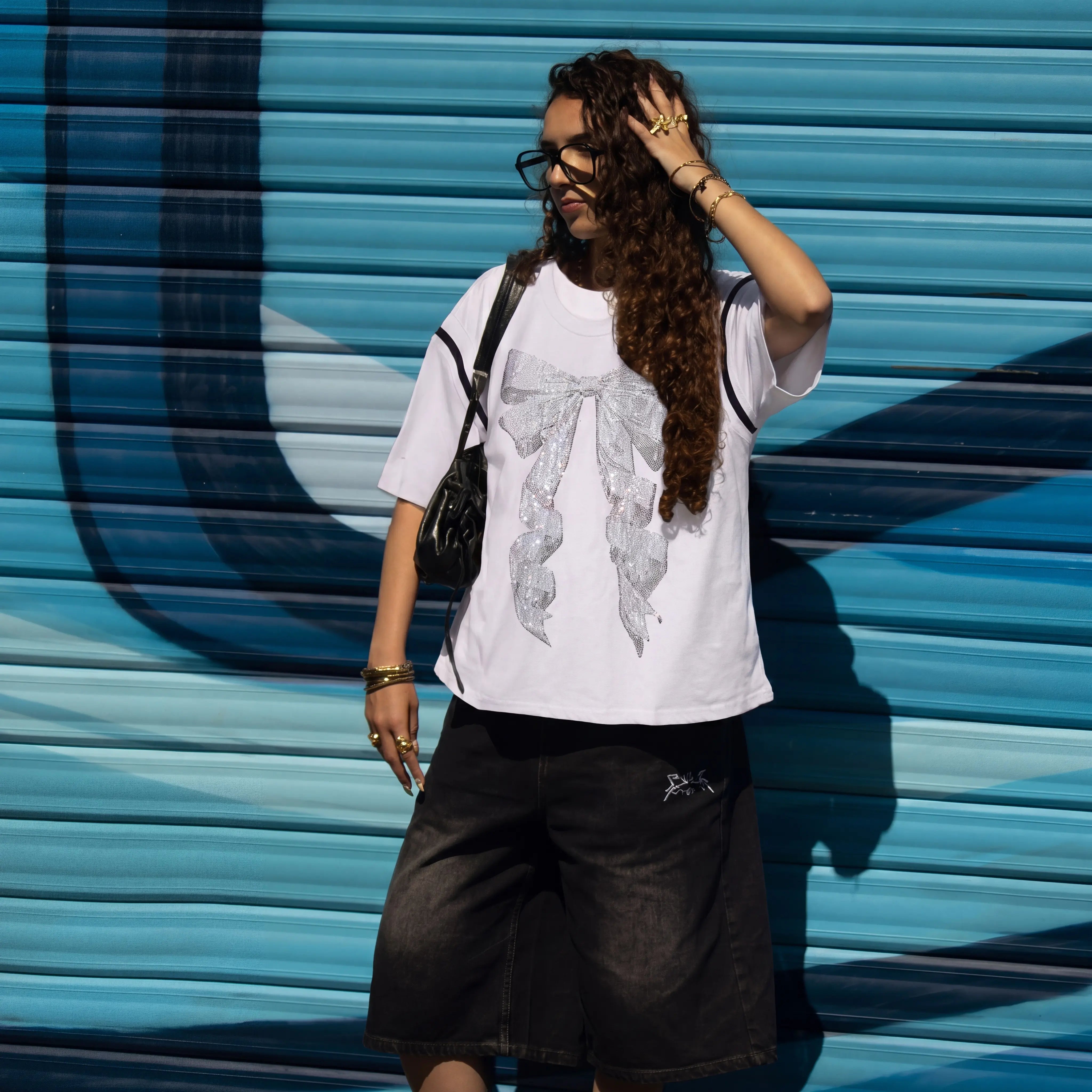 Person wearing a white t-shirt with a graphic design and black shorts against a blue wall. TCH Rhinestone Bow White T-Shirt