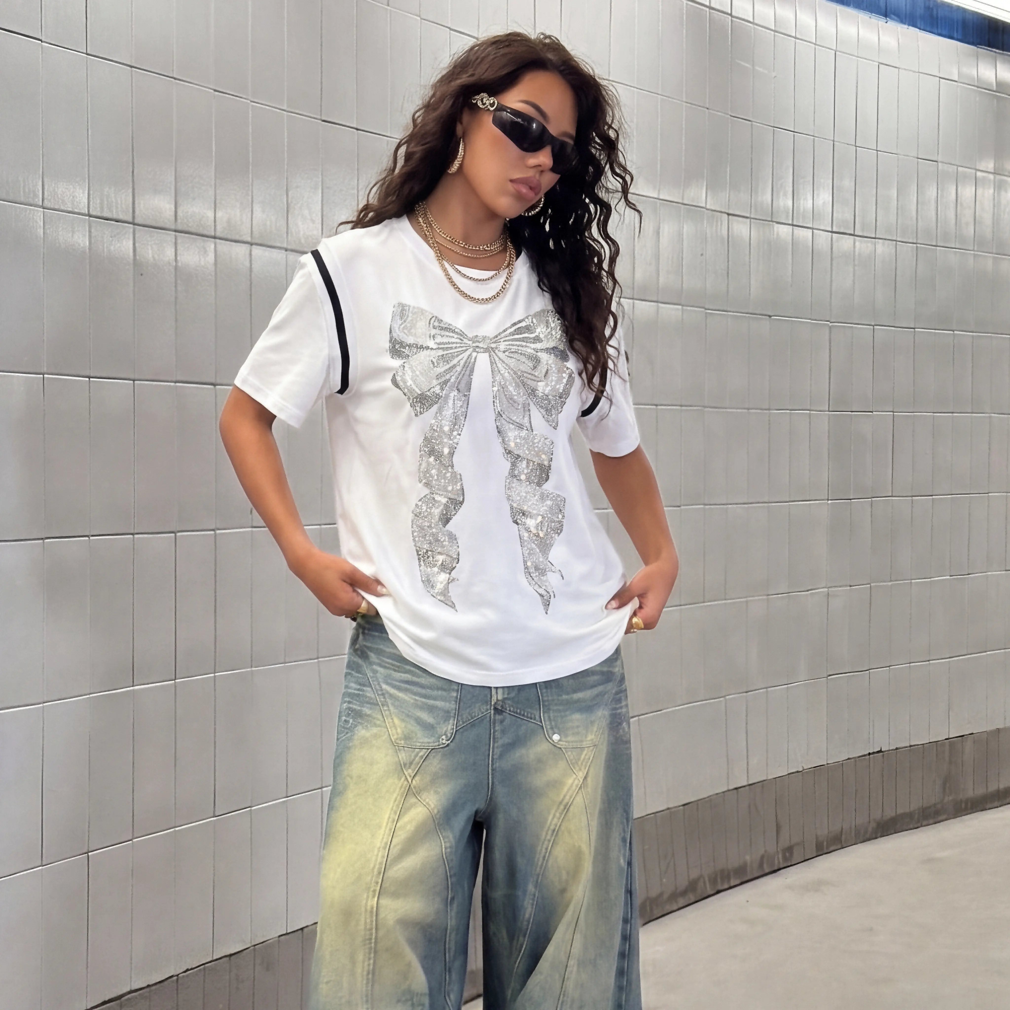 Woman wearing a white t-shirt with a large bow design and light blue jeans against a tiled wall. TCH Rhinestone Bow White T-Shirt