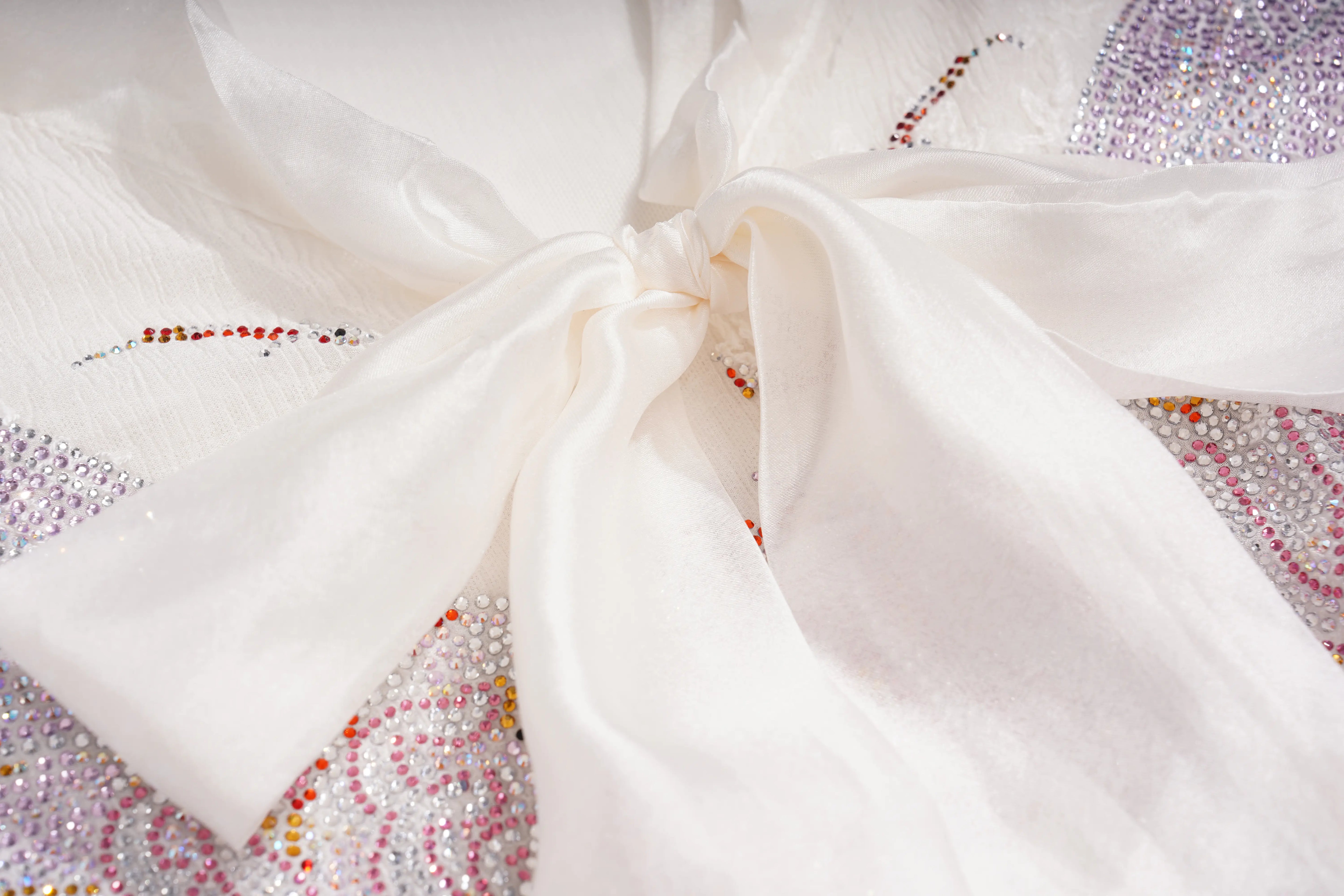 A close-up of a white sheer fabric bow tied in the center, layered over a sparkling embellished surface with silver, pink, and red rhinestones. TCH Rhinestone Butterfly Bow Lace Blouse