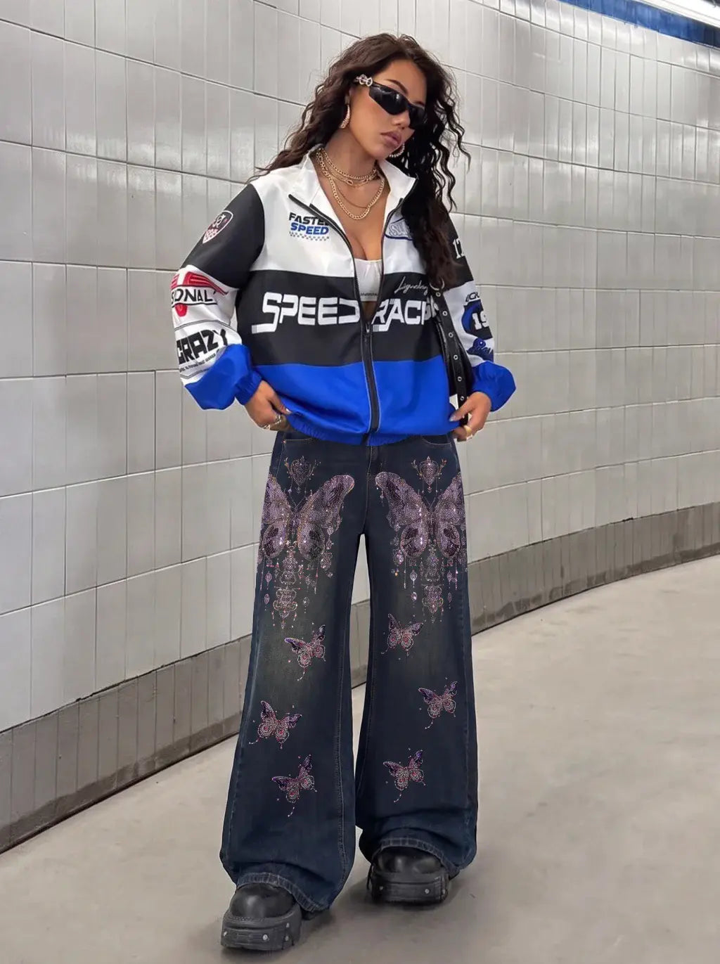 A woman stands in a tiled tunnel wearing a racing-inspired zip-up jacket with 'SPEED RACING' across the chest, blue and white panels, and sponsor-style graphics, paired with dark wide-leg jeans featuring a purple butterfly print and black boots. TCH Rhinestone Butterfly Charm Wide-Leg Denim Jeans