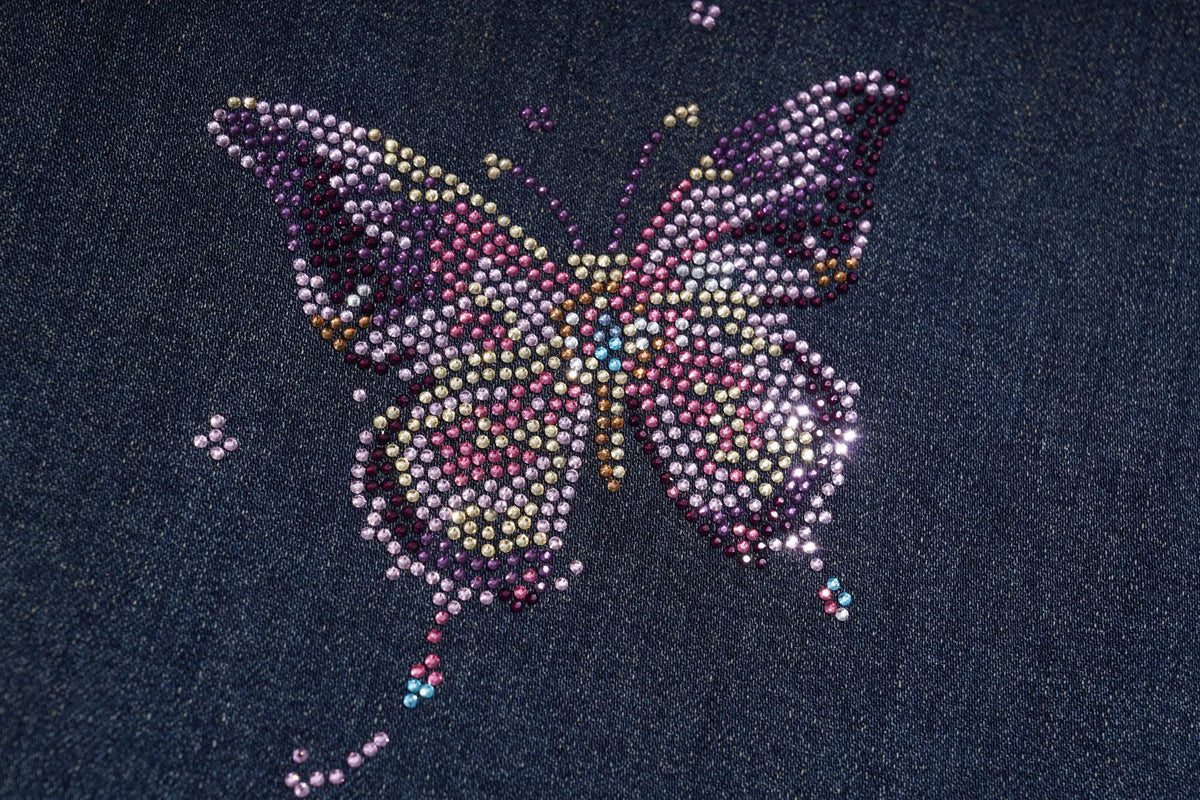 A butterfly-shaped rhinestone or bead design on a dark navy blue fabric background. TCH Rhinestone Butterfly Charm Wide-Leg Denim Jeans