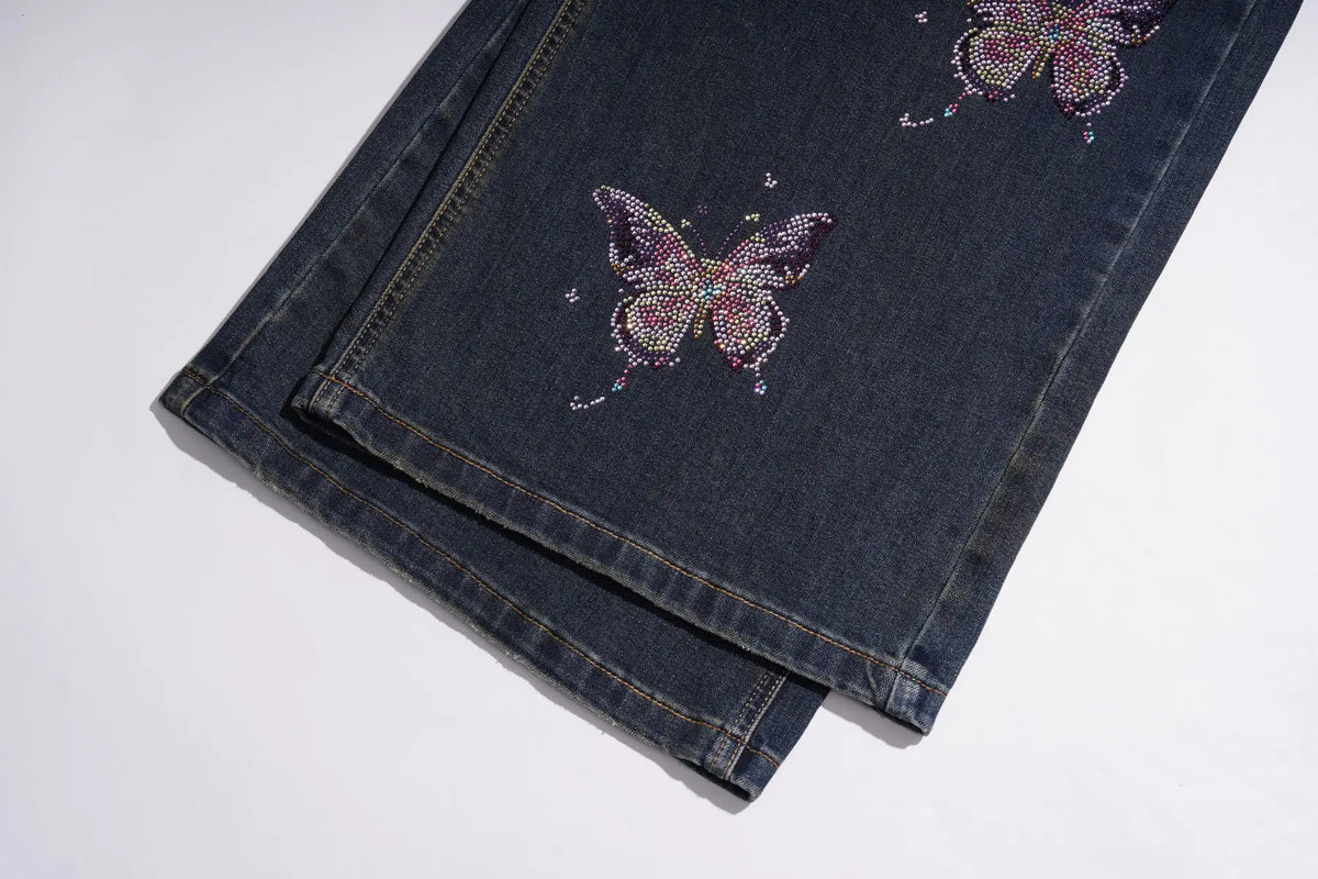 Dark blue denim jeans folded on a white background, featuring two large butterfly designs embellished with pink, purple, and silver rhinestones. TCH Rhinestone Butterfly Charm Wide-Leg Denim Jeans