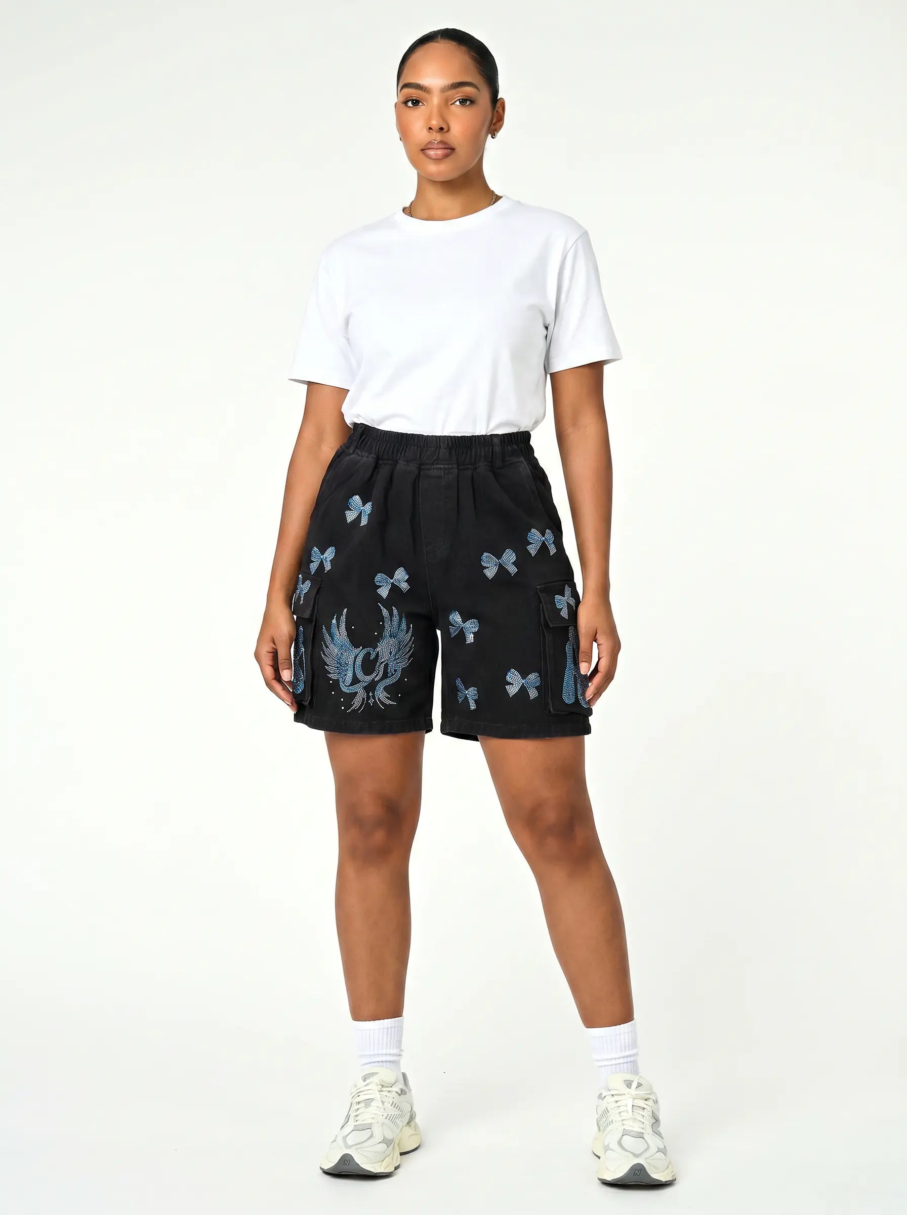 A model stands against a plain white studio background wearing a white t-shirt, black shorts with blue dragon and butterfly prints, white socks, and white sneakers. TCH Rhinestone Butterfly & Dragon Graphic Black Cargo Shorts