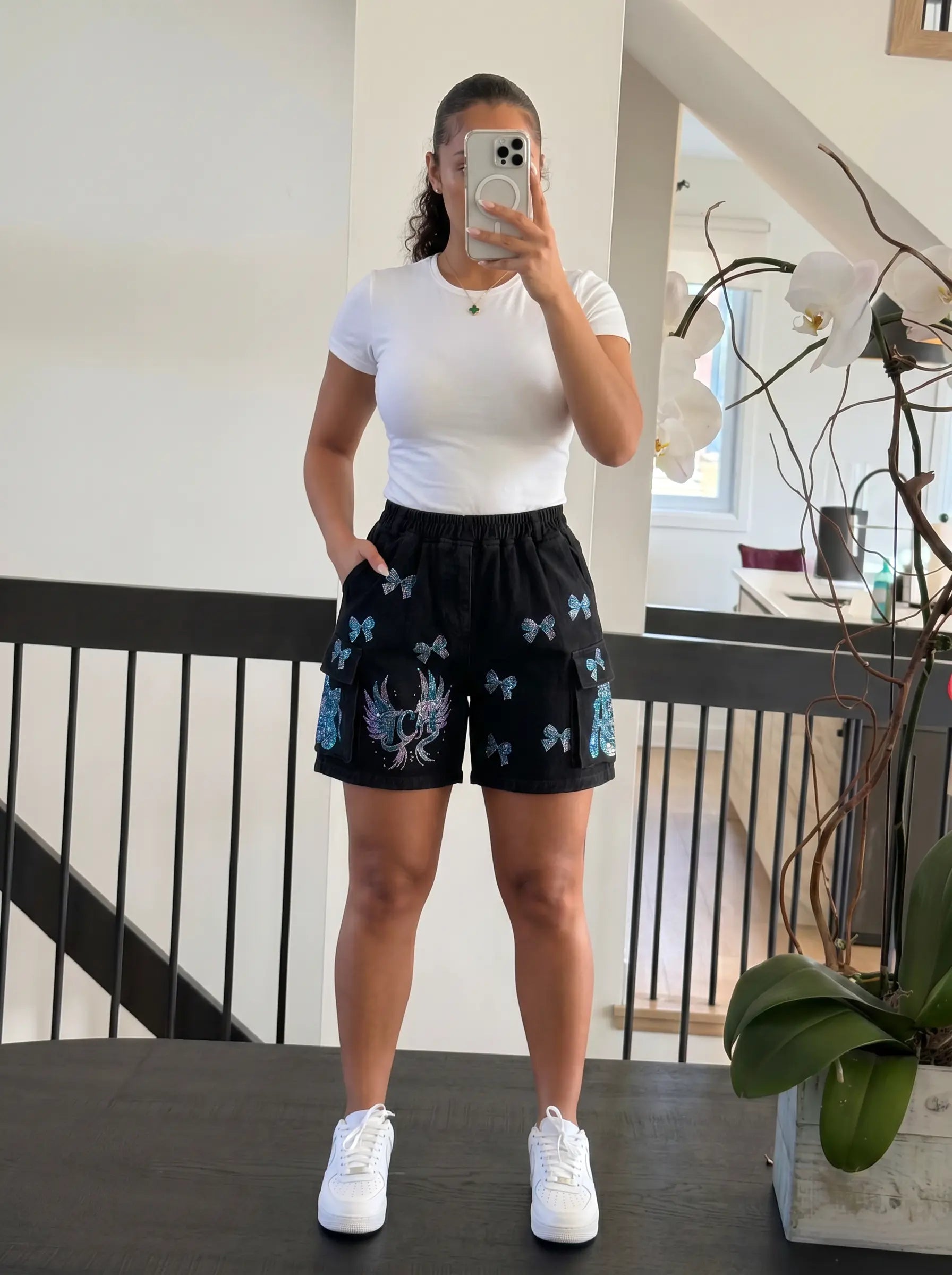 A woman takes a mirror selfie wearing black athletic shorts with blue butterfly and white eagle graphics, paired with a white t-shirt and white sneakers, in an indoor setting. TCH Rhinestone Butterfly & Dragon Graphic Black Cargo Shorts