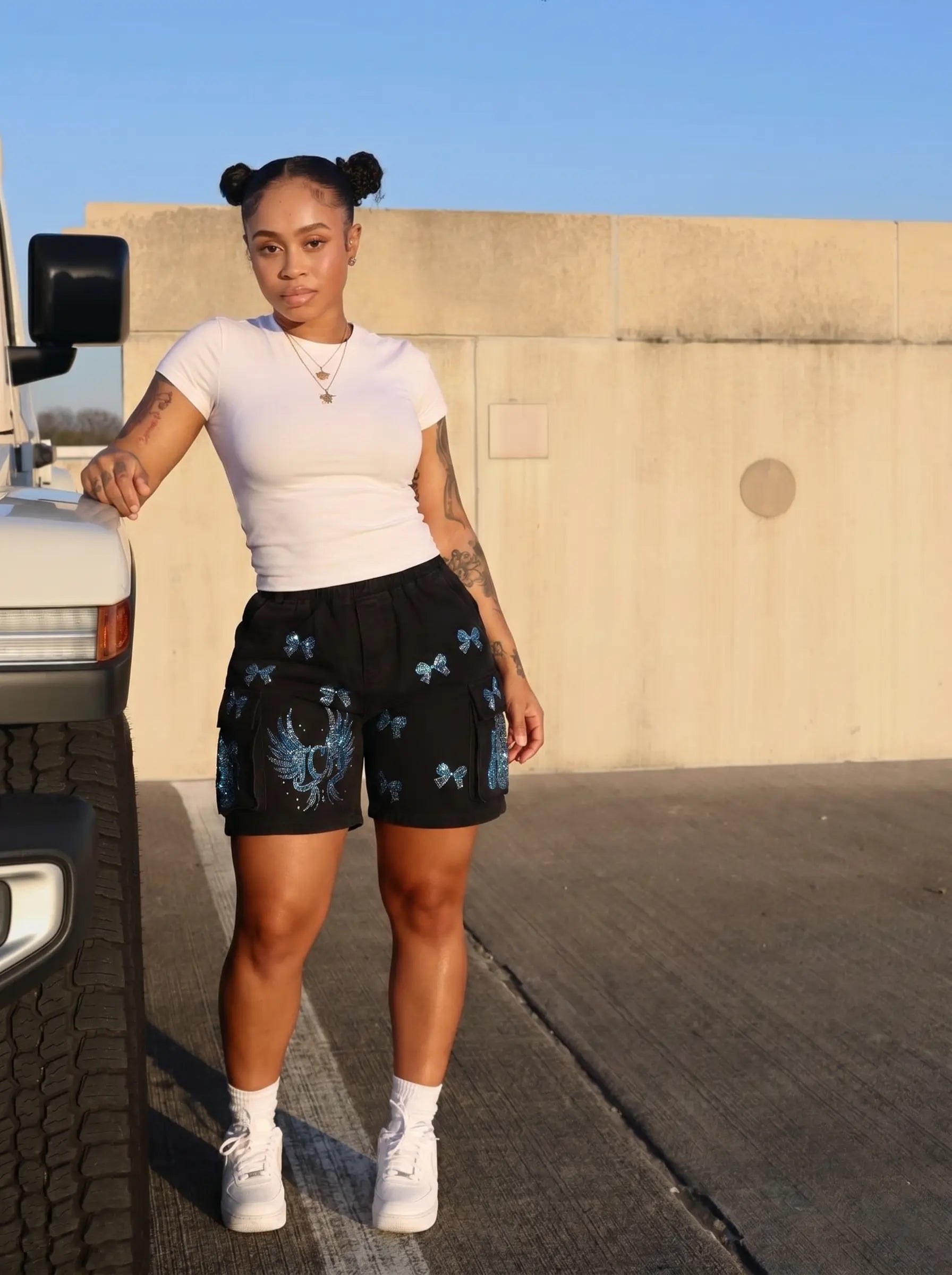 A woman stands outdoors next to a vehicle wearing a white fitted t-shirt and black athletic shorts with blue butterfly and bird graphics. She is posing in front of a concrete wall under a clear blue sky. TCH Rhinestone Butterfly & Dragon Graphic Black Cargo Shorts