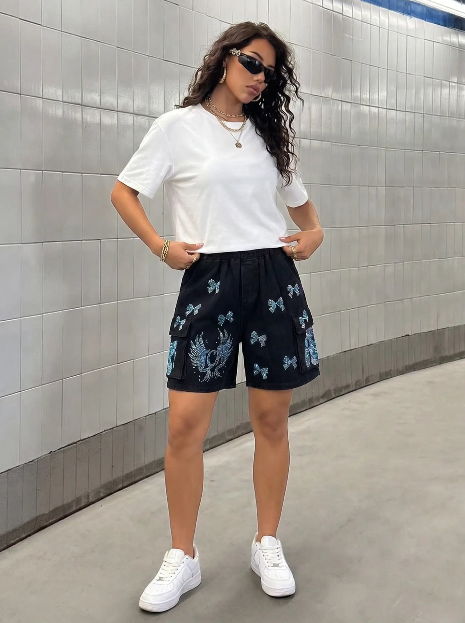 A woman wearing a white t-shirt and black cargo shorts with blue butterfly prints, standing in a tiled tunnel. TCH Rhinestone Butterfly & Dragon Graphic Black Cargo Shorts