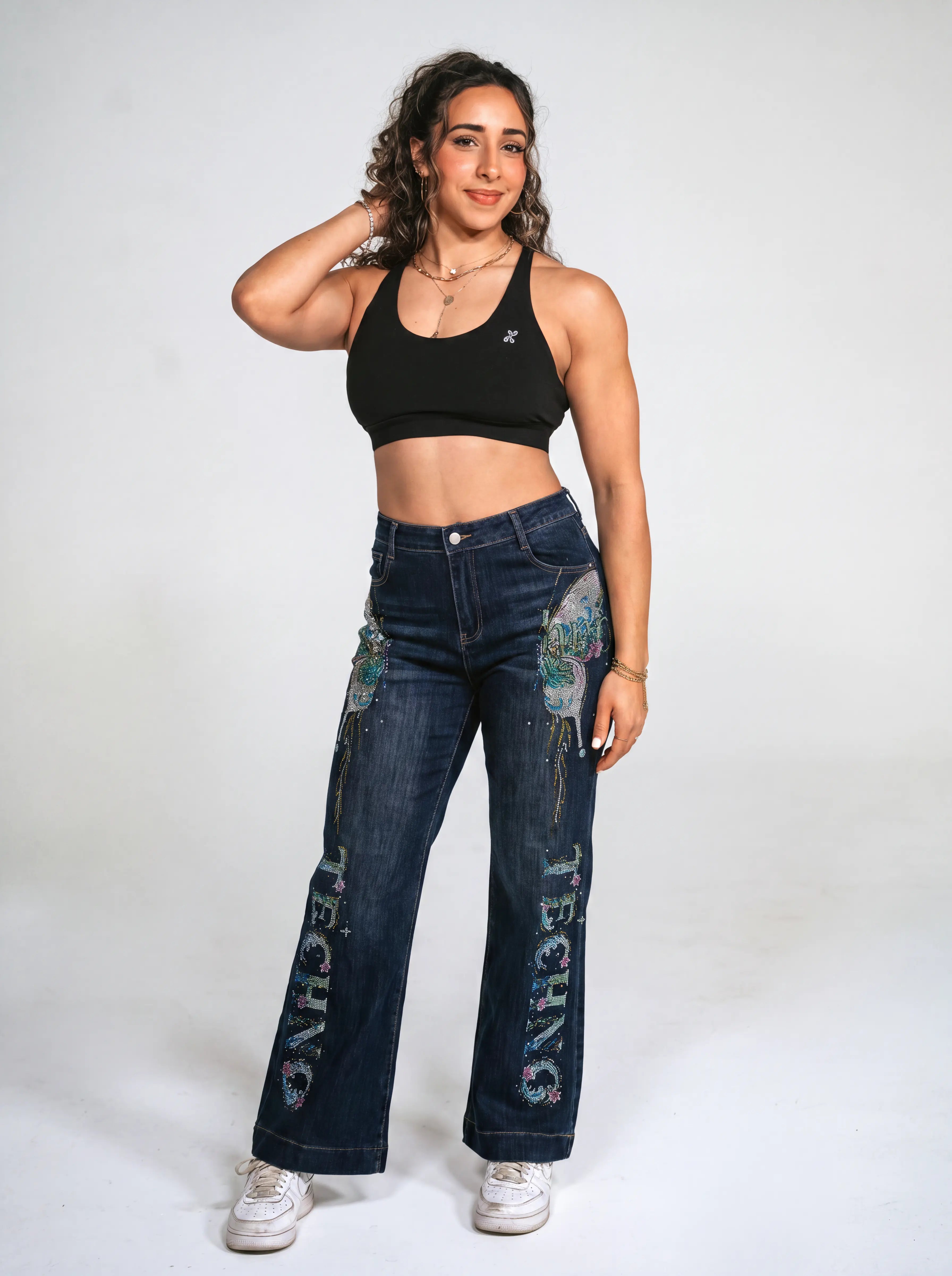 A woman modeling a pair of dark blue wide-leg jeans with colorful graphic prints and 'TECHNO' text down the front, paired with a black sports bra and white sneakers, against a plain white studio background. TCH Rhinestone Butterfly Graphic Wide-Leg Jeans