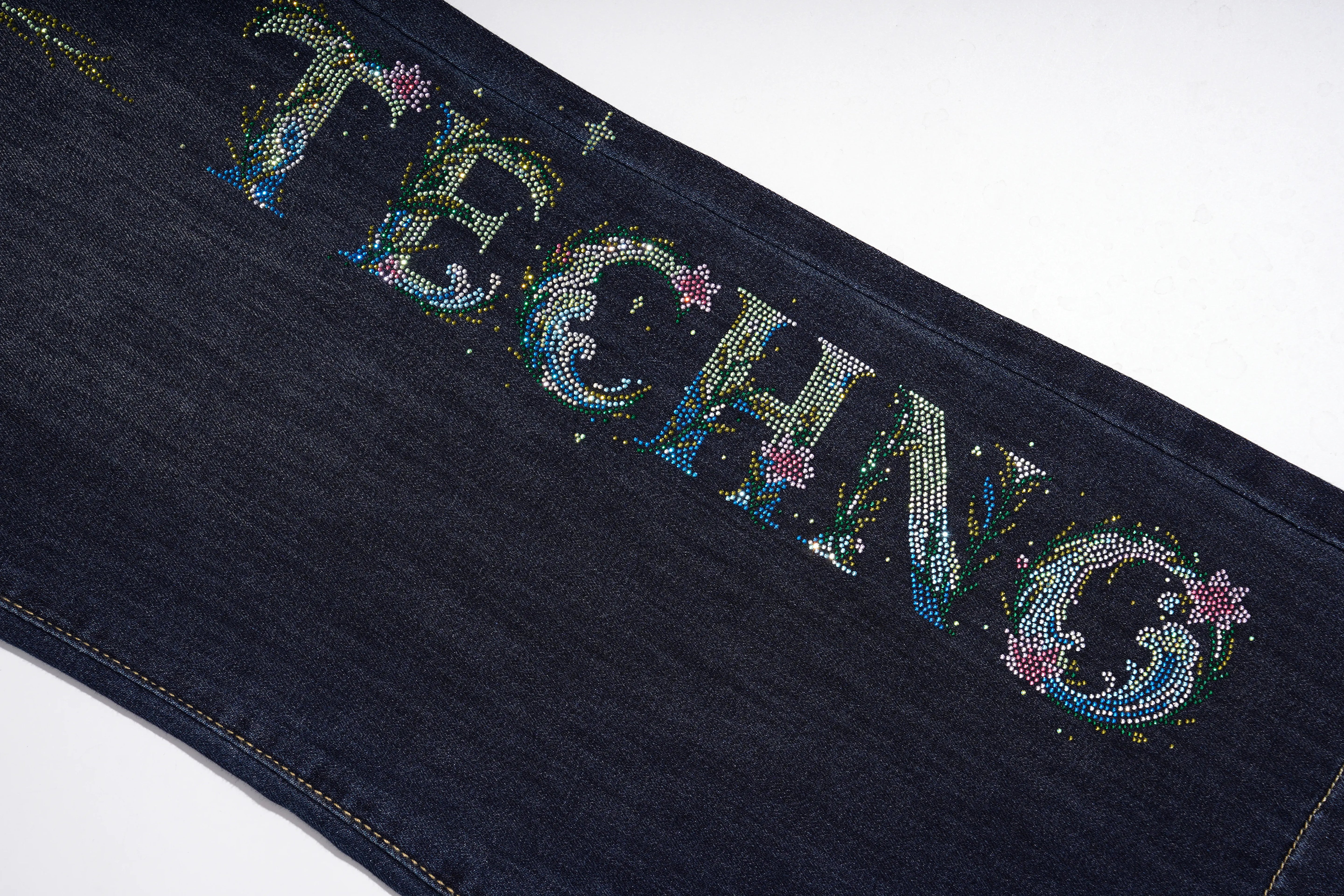 Close-up of a dark blue denim garment featuring the word 'TECHNO' spelled out in large, embellished letters with rhinestones and small pink floral accents, set against a plain white background. TCH Rhinestone Butterfly Graphic Wide-Leg Jeans