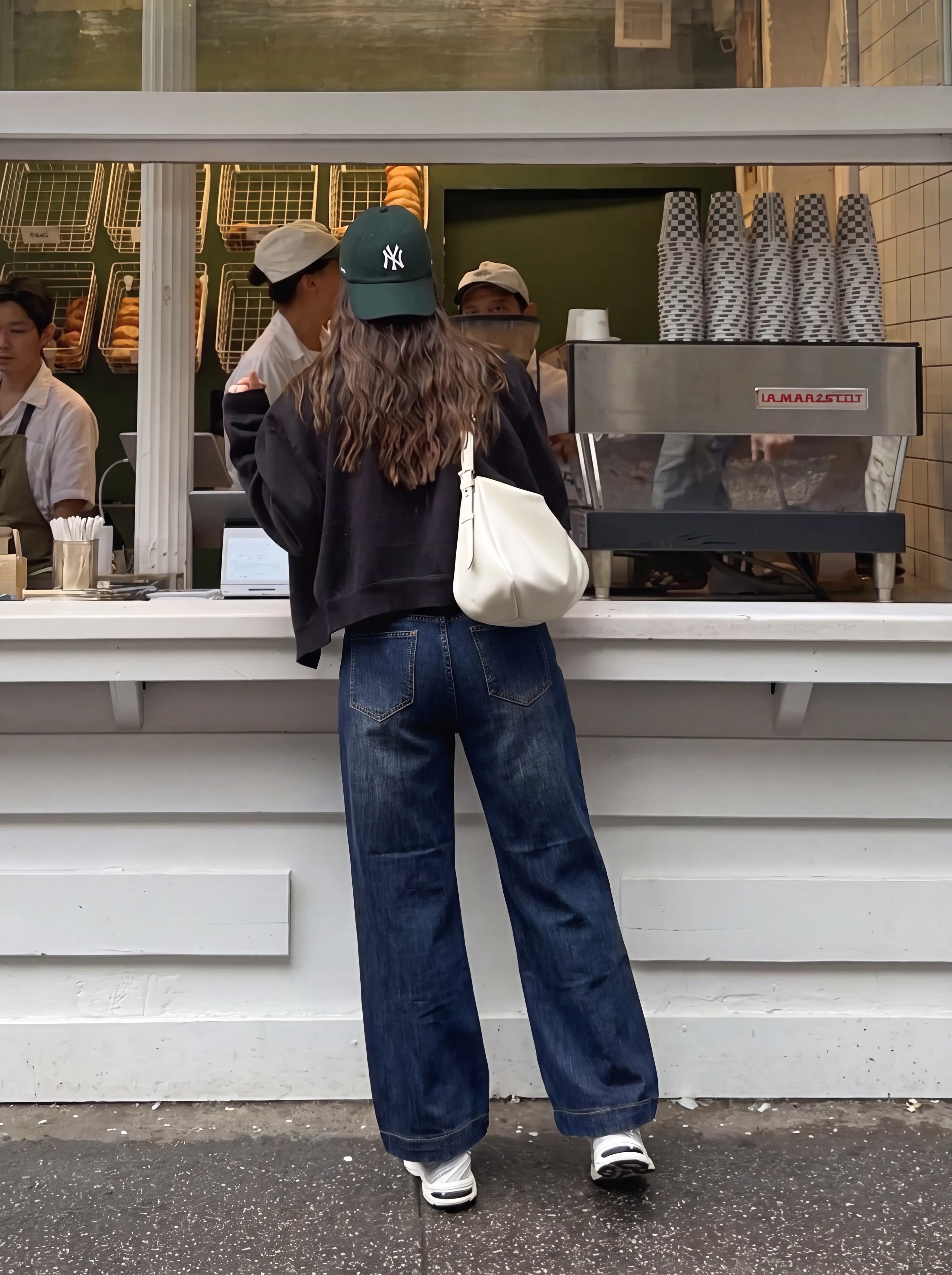 A person standing at a cafe counter, viewed from behind, wearing a dark navy cropped sweatshirt, wide-leg dark blue jeans, a green baseball cap, and carrying a white shoulder bag. TCH Rhinestone Butterfly Graphic Wide-Leg Jeans