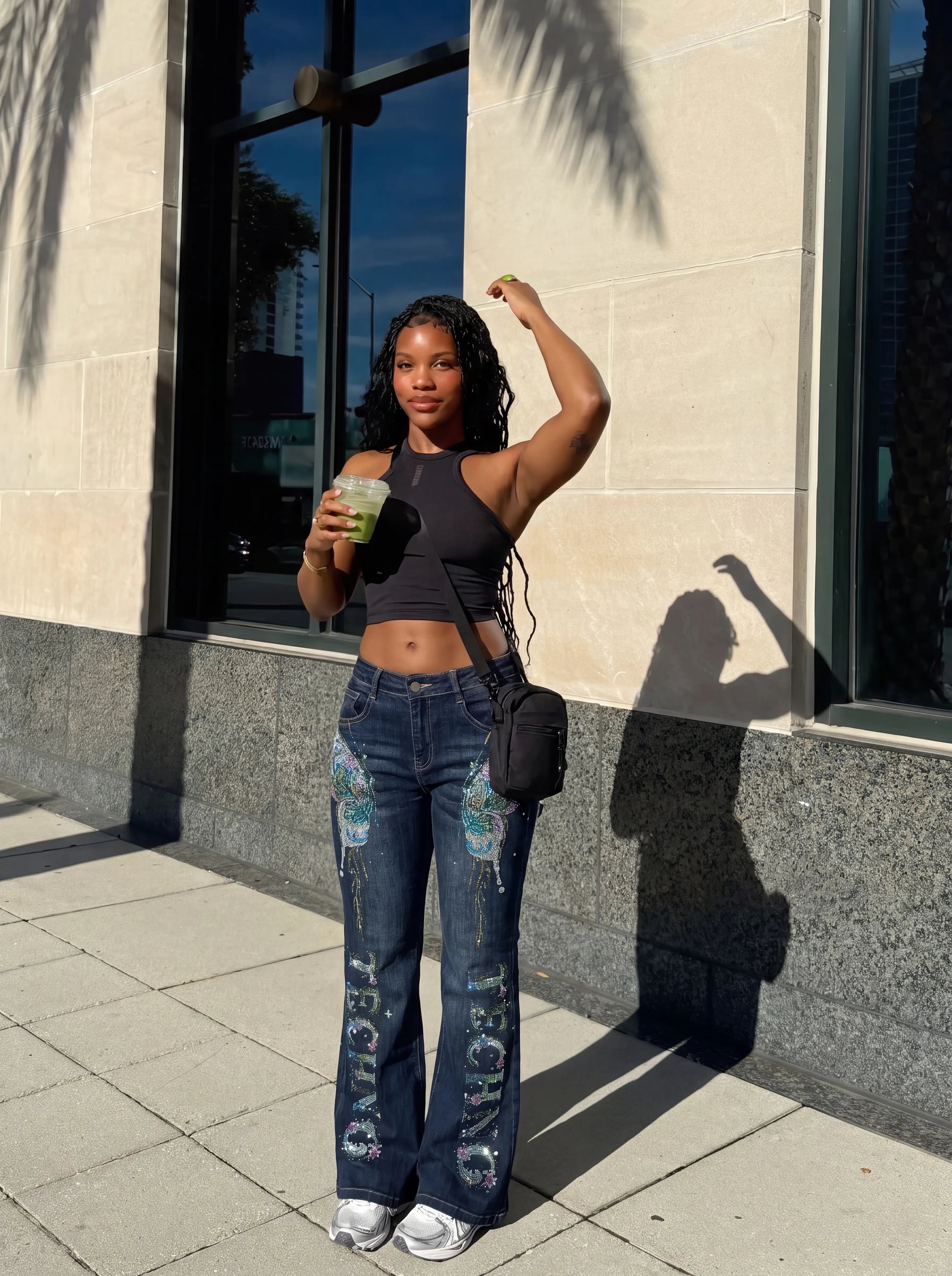 A woman stands outdoors on a sidewalk in front of a stone building, wearing a black ribbed crop top, distressed blue flare jeans with colorful graphic patches, and white sneakers. She holds a green drink and carries a small black shoulder bag. TCH Rhinestone Butterfly Graphic Wide-Leg Jeans