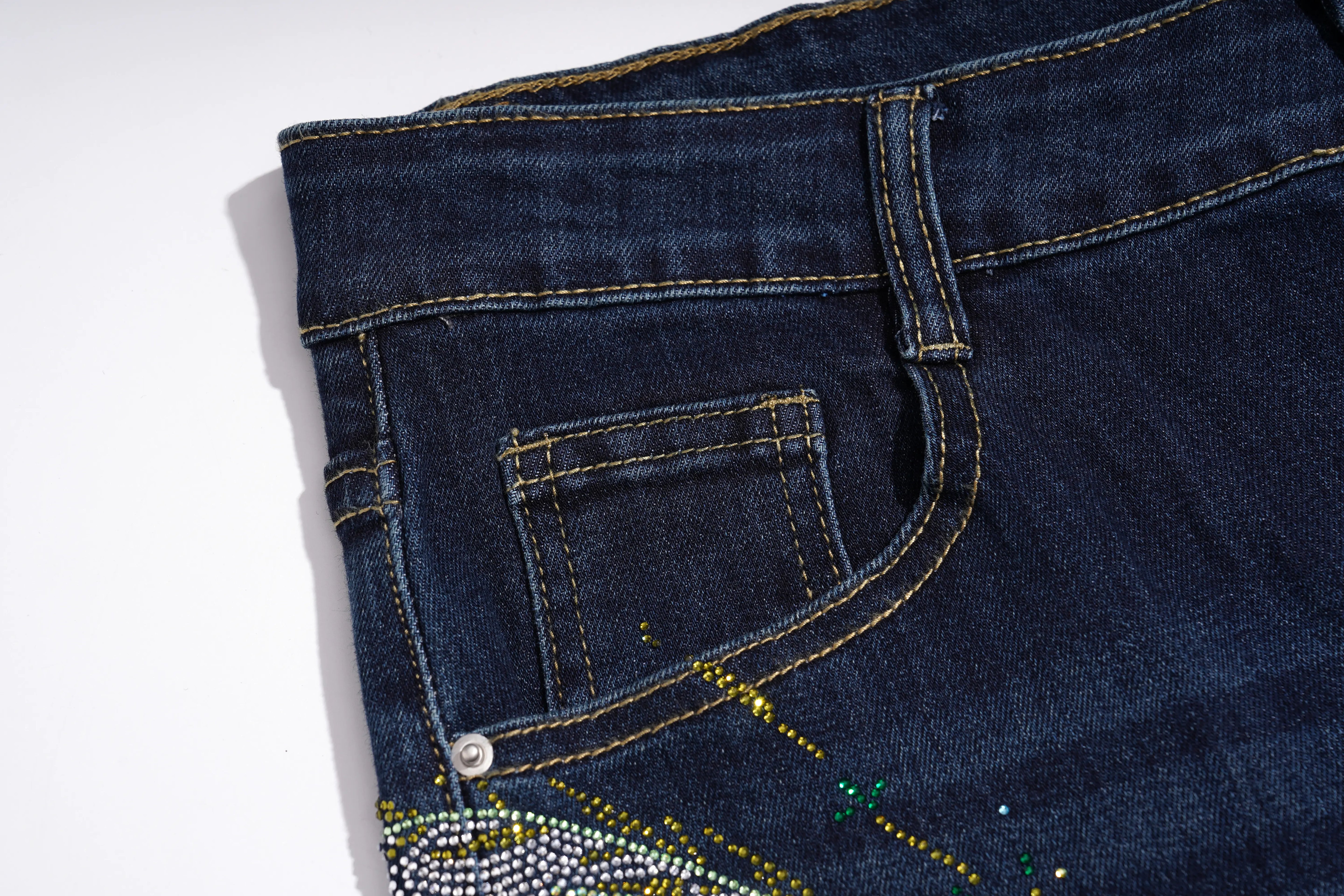 Close-up of the back pocket area of dark blue denim jeans with gold contrast stitching and a decorative embellishment near the bottom corner. TCH Rhinestone Butterfly Graphic Wide-Leg Jeans