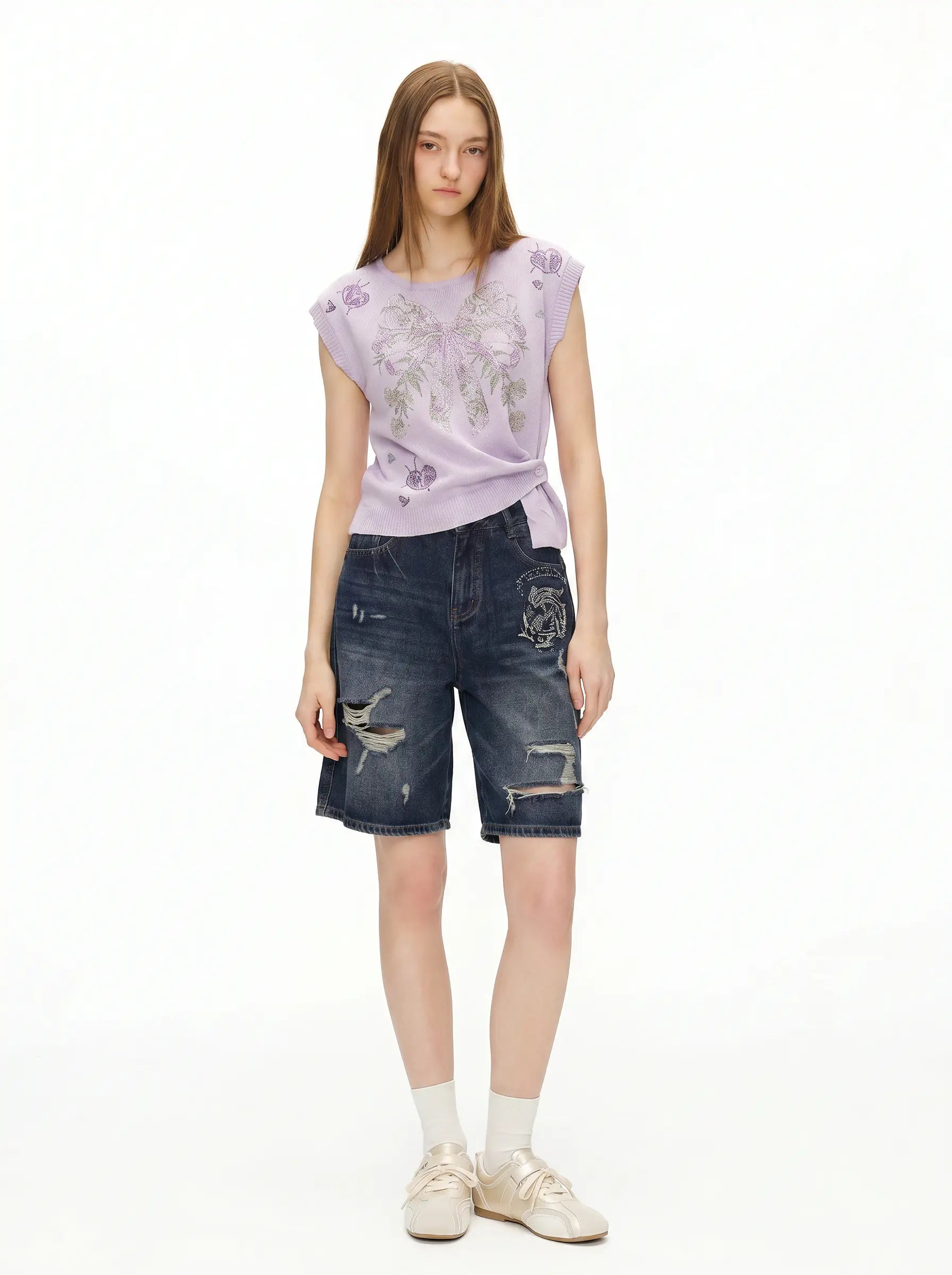 A model stands against a white studio background wearing a lavender sleeveless top with a floral print, dark blue distressed denim shorts, white socks, and beige sneakers. TCH Rhinestone Butterfly Knit Twist Top