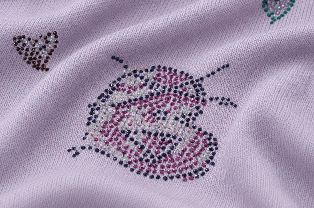 Close-up of a light lavender knit fabric featuring a decorative ladybug design made from small rhinestones in pink, white, and dark gray, with small heart motifs in the corners. TCH Rhinestone Butterfly Knit Twist Top