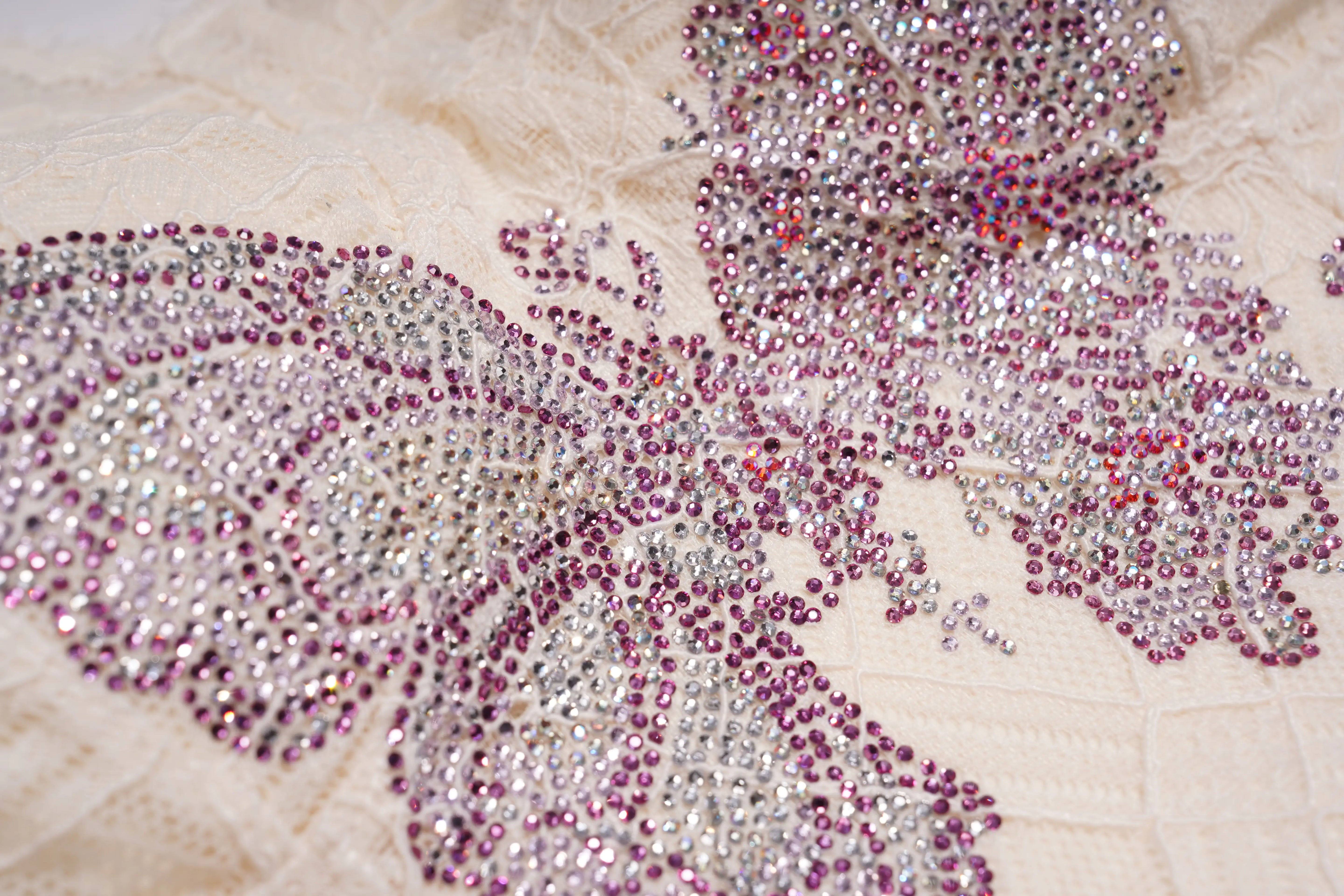 A close-up of a butterfly-shaped embellishment covered in sparkling rhinestones and beads, featuring silver, pink, and purple tones on a textured cream lace background. TCH Rhinestone Butterfly Lace Cami Top