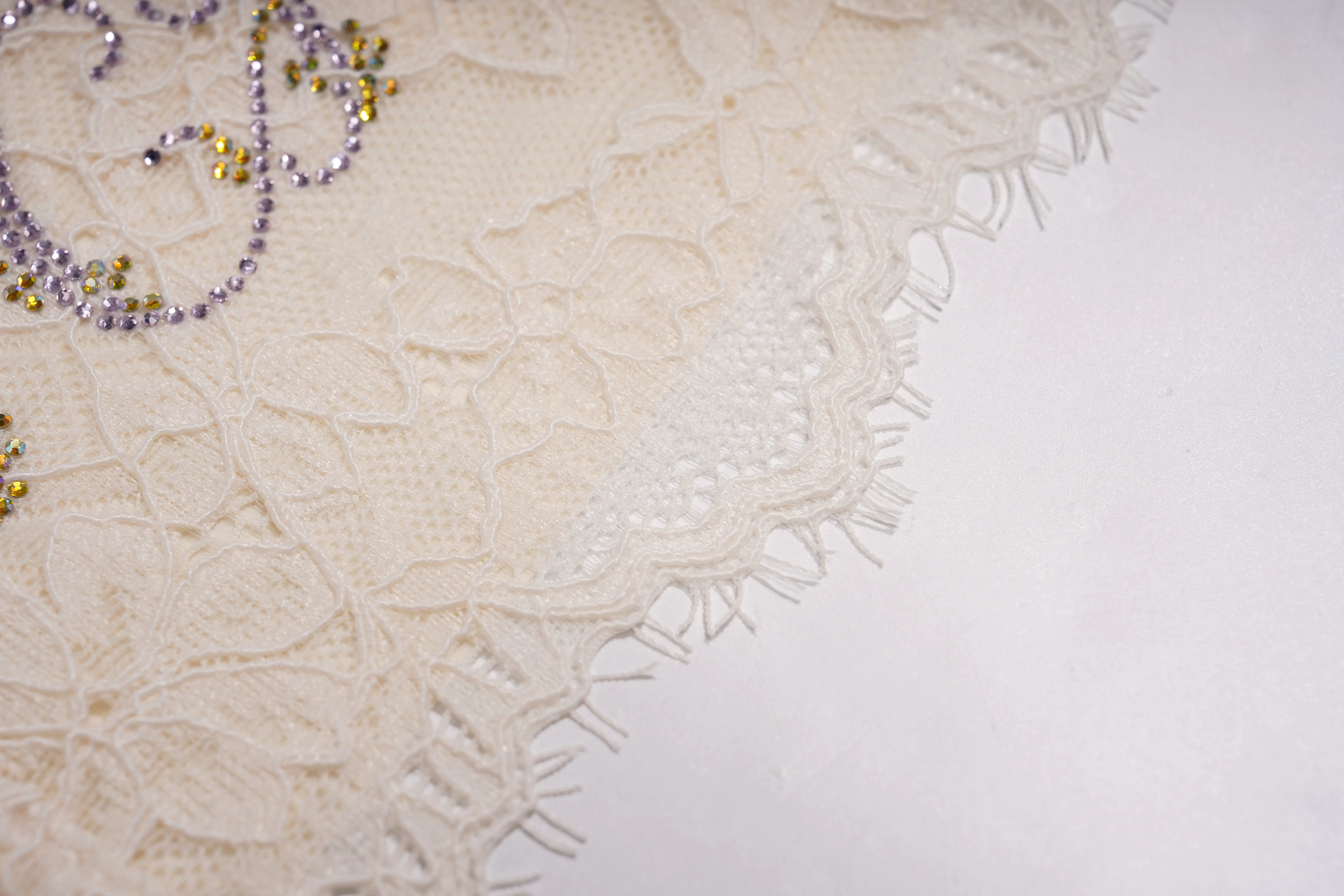 Close-up of an off-white lace textile with floral embroidery and scalloped fringe, decorated with small purple and yellow beaded embellishments on a plain white background. TCH Rhinestone Butterfly Lace Cami Top