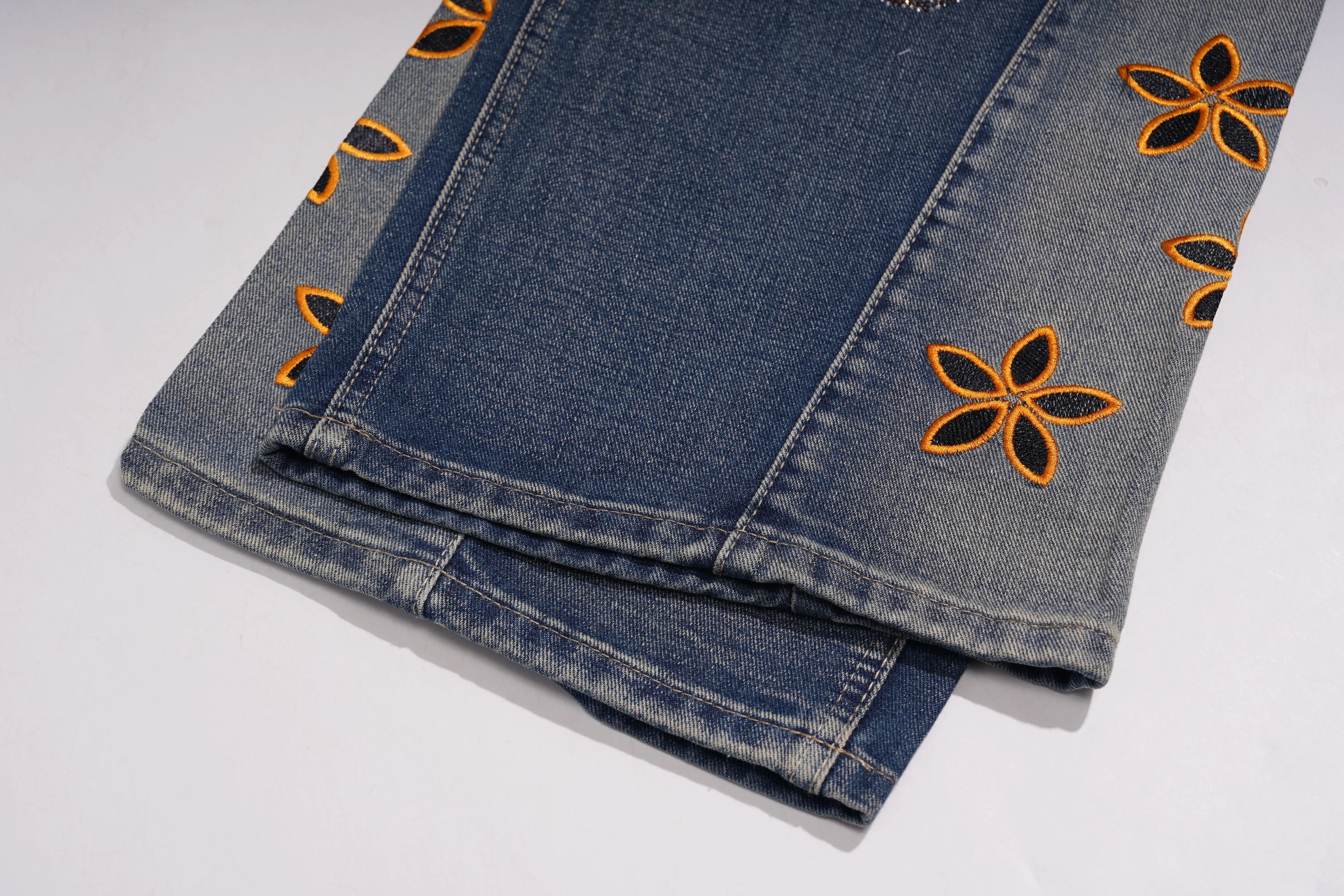 Folded blue denim jeans with orange floral embroidery on a white background, TCH Rhinestone Butterfly & Lily Distressed Denim Jeans