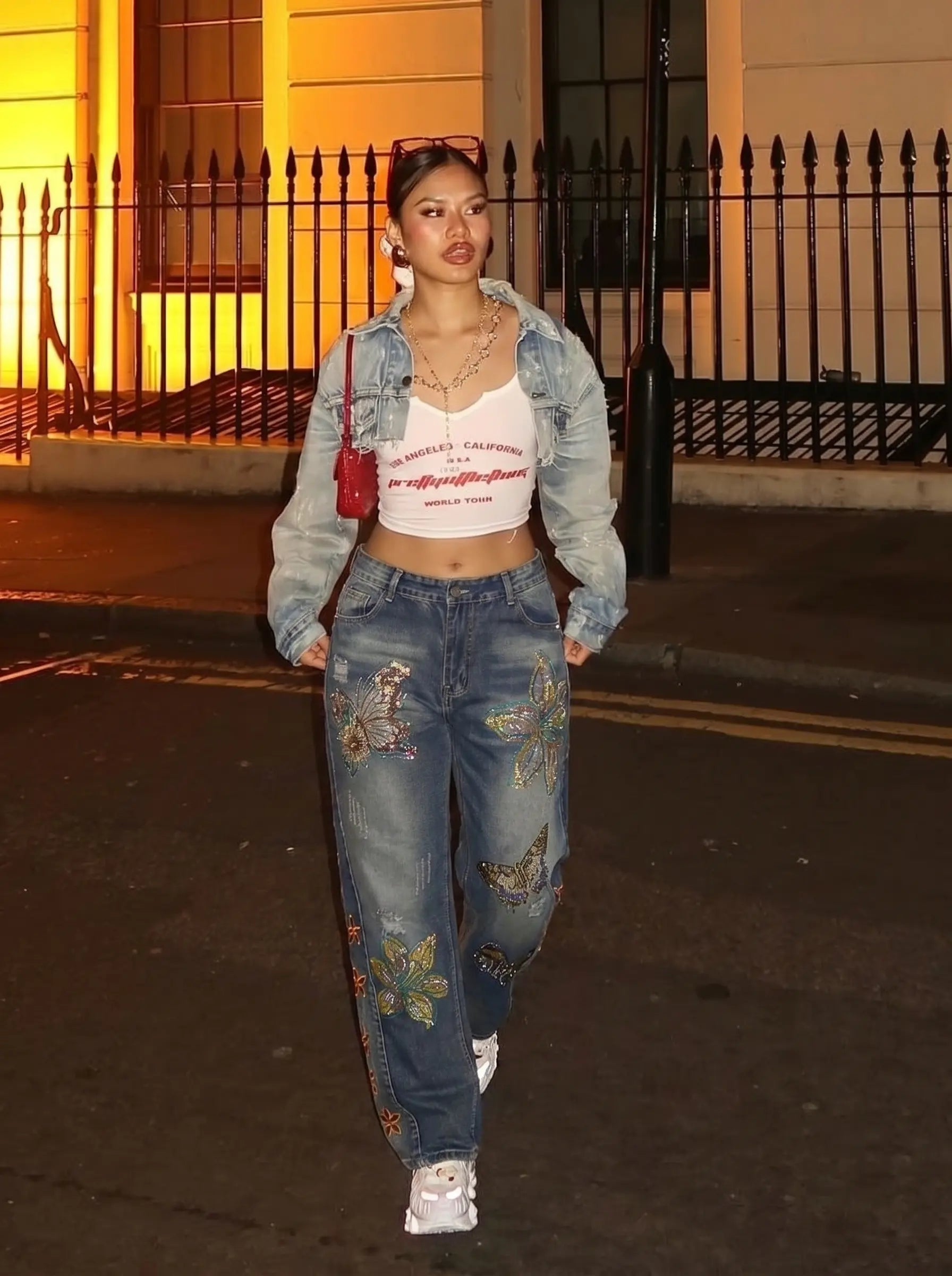 Woman walking on a street at night wearing a denim jacket, white crop top, and embroidered jeans. TCH Rhinestone Butterfly & Lily Distressed Denim Jeans