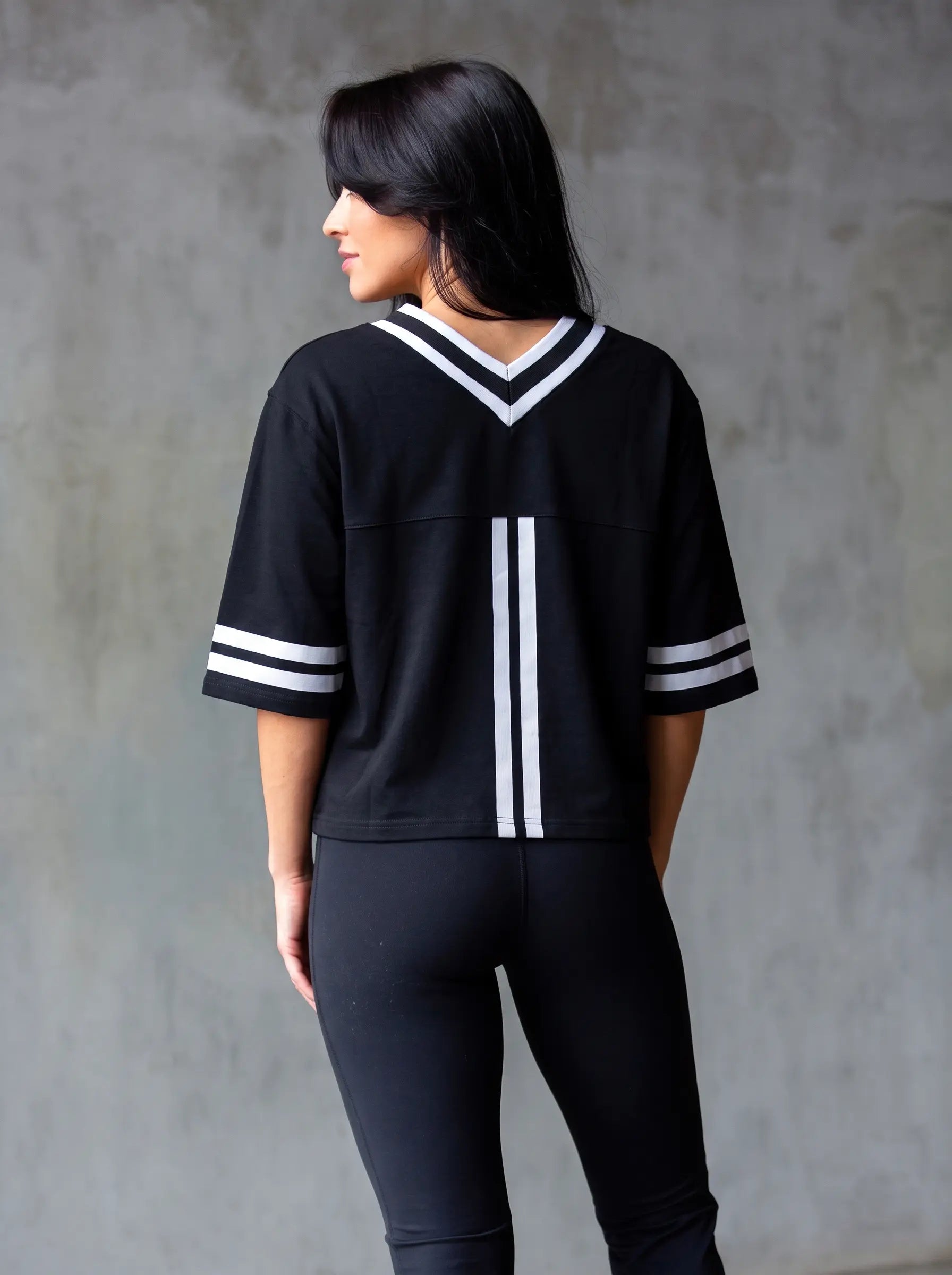 A woman shown from the back wearing a black short-sleeve athletic-style top with white V-neck trim and white stripes on the sleeves and center back, paired with black leggings, against a textured gray wall. TCH Rhinestone Butterfly V-Neck Oversized Baseball T-Shirt