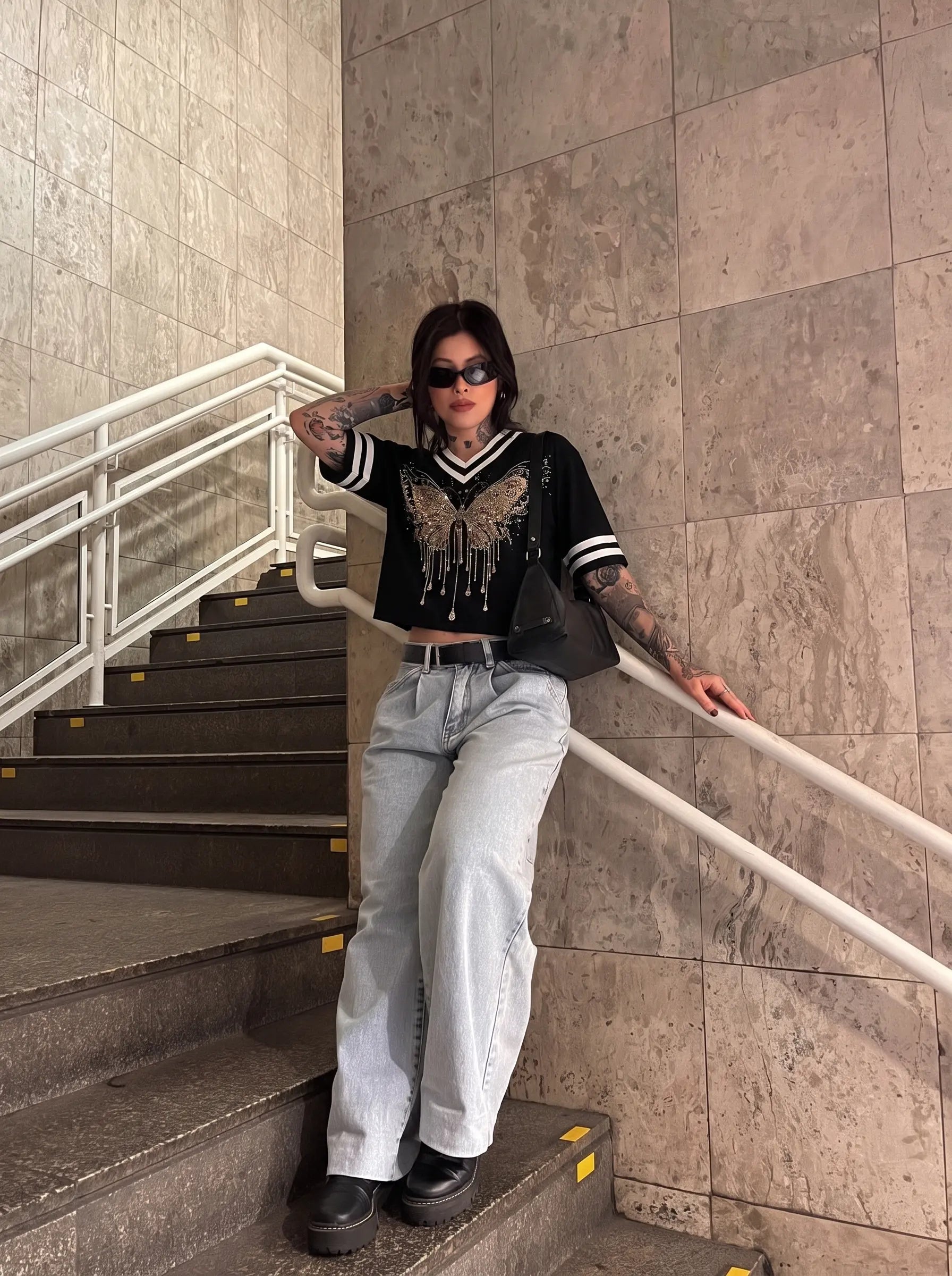 A person posing on an indoor staircase wearing a black V-neck crop top with a metallic butterfly graphic and white striped sleeves, paired with light-wash wide-leg jeans and black boots. TCH Rhinestone Butterfly V-Neck Oversized Baseball T-Shirt
