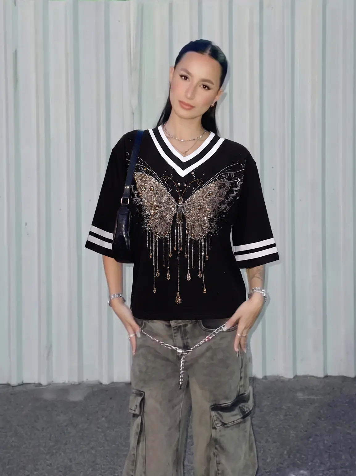 A woman stands against a light gray corrugated metal wall wearing a black short-sleeve V-neck top with white stripes on the sleeves and a large metallic butterfly embellishment with dangling fringe across the chest. She is also wearing gray cargo pants and carrying a black shoulder bag. TCH Rhinestone Butterfly V-Neck Oversized Baseball T-Shirt