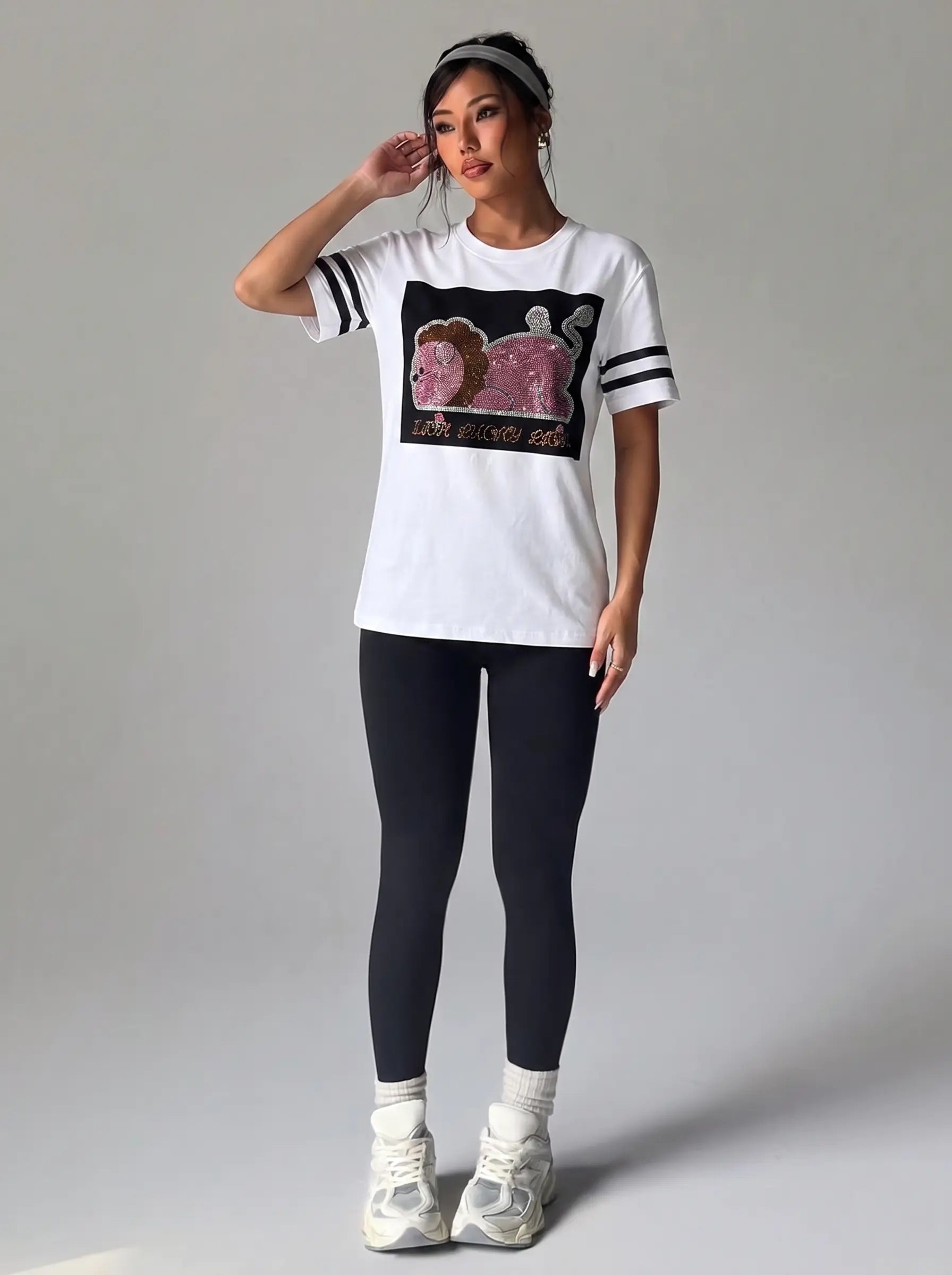 A woman modeling a white short-sleeve athletic T-shirt with black sleeve stripes and a large black chest graphic featuring pink and brown cartoon-style figures. She is wearing black leggings, white socks, and white sneakers against a plain light gray studio background. TCH Rhinestone Cartoon Pig Graphic Baseball T-Shirt