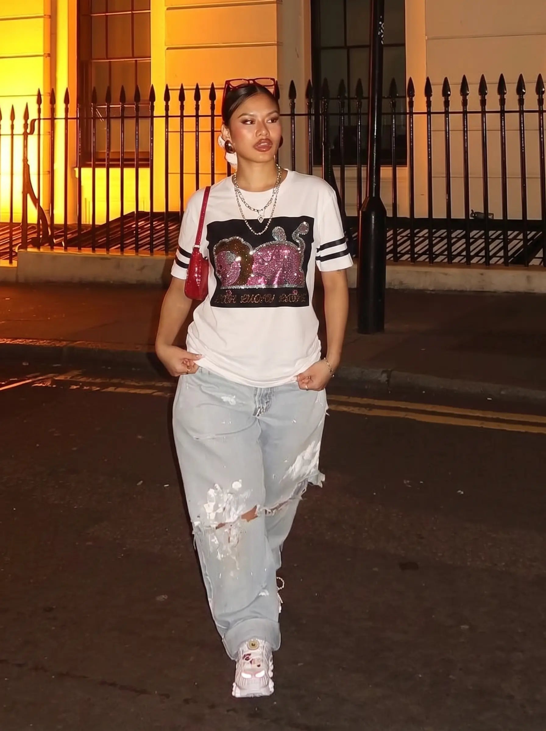 A woman walking outdoors at night wearing a white graphic t-shirt with a pink sequined elephant design, light-wash distressed jeans, and white sneakers. TCH Rhinestone Cartoon Pig Graphic Baseball T-Shirt