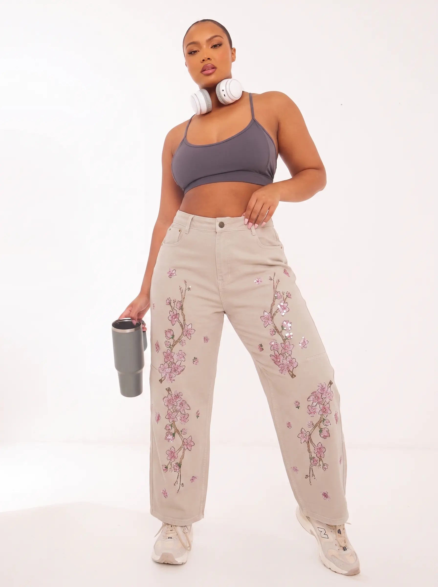 A model wearing beige wide-leg jeans with pink floral embroidery, a gray sports bra, and white sneakers, holding a gray tumbler against a white studio background. TCH Rhinestone Cherry Blossom Straight-Leg Jeans