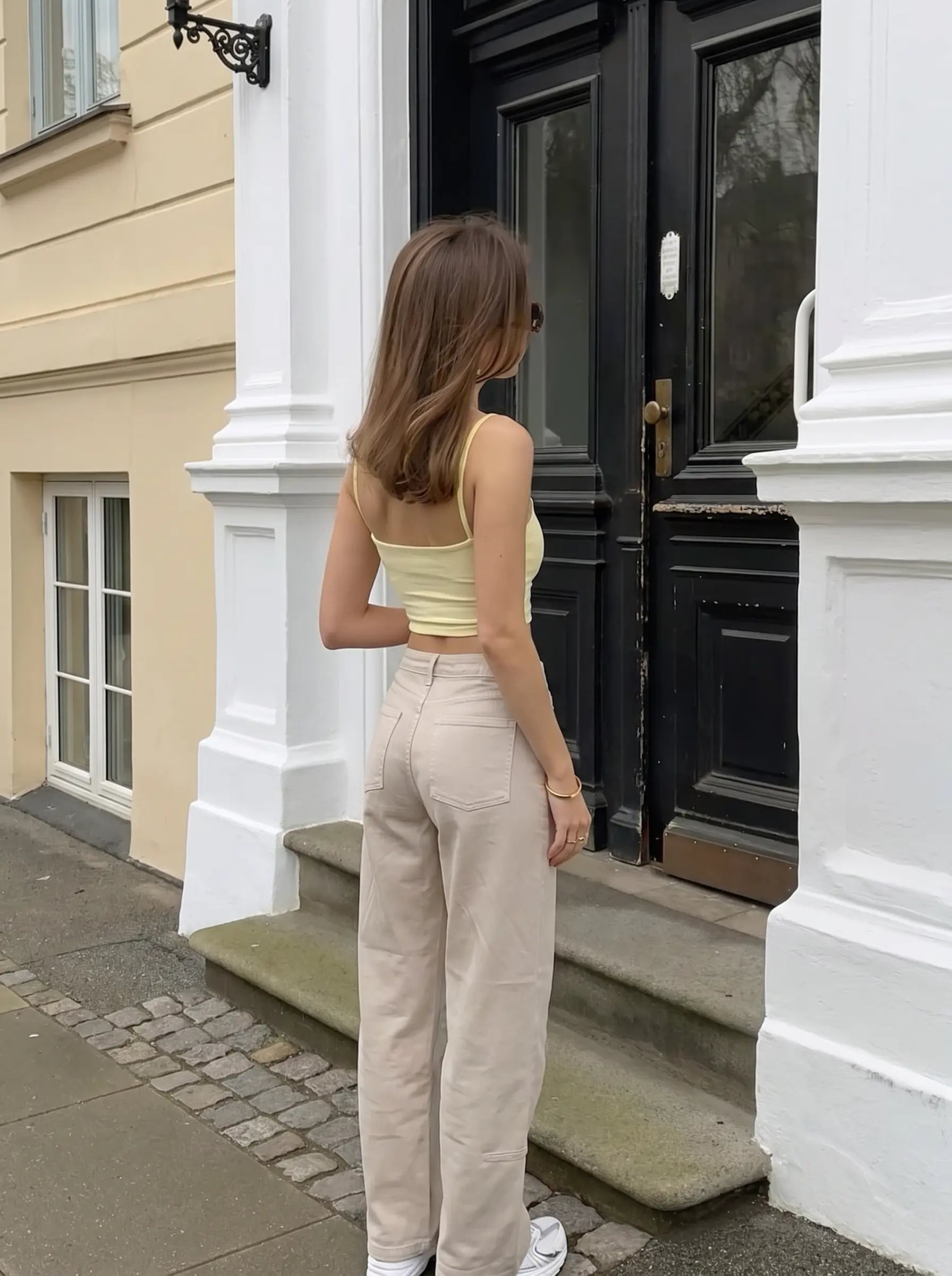 A woman stands with her back to the camera in front of a black door and white columns, wearing a pale yellow ribbed tank top and light beige wide-leg trousers. TCH Rhinestone Cherry Blossom Straight-Leg Jeans