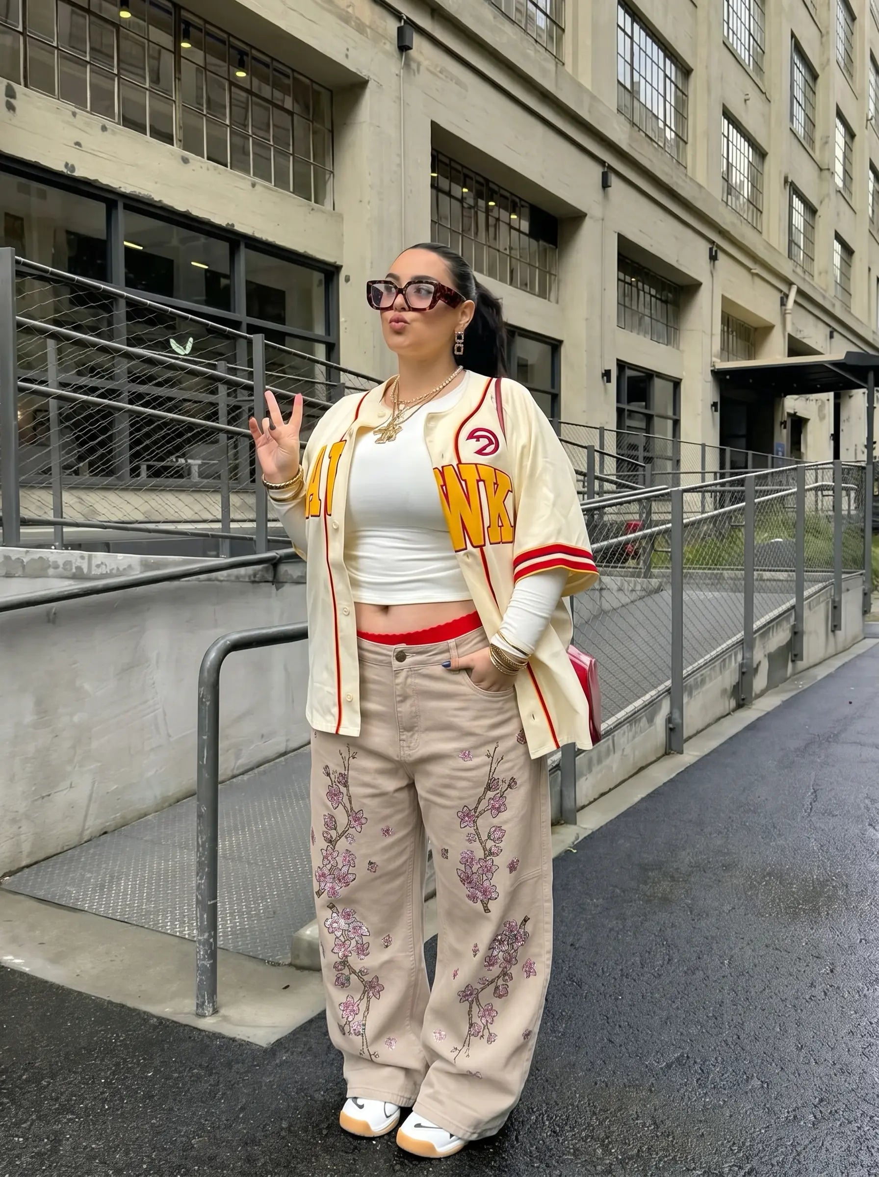 A woman stands outdoors on a city street in front of a concrete building, wearing a cream varsity-style jacket with red and yellow lettering, a white crop top, beige wide-leg pants with purple floral embroidery, and white sneakers. TCH Rhinestone Cherry Blossom Straight-Leg Jeans