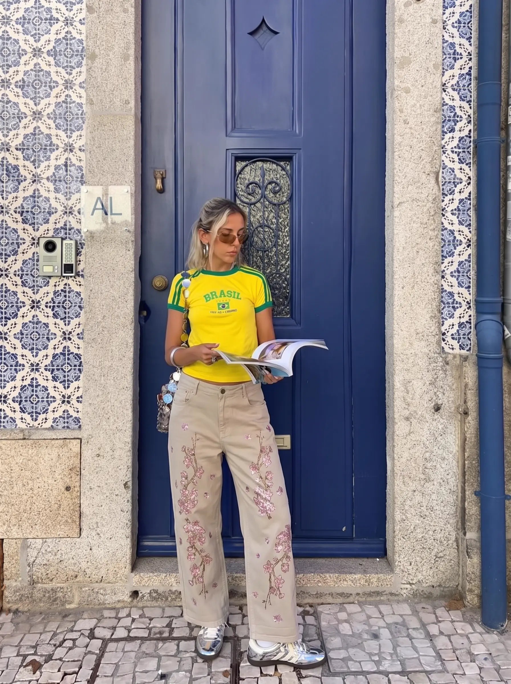 A woman stands in front of a blue door wearing a bright yellow t-shirt with green trim and 'BRASIL' printed on the chest, paired with beige wide-leg pants featuring pink floral embroidery. She is holding a magazine. TCH Rhinestone Cherry Blossom Straight-Leg Jeans