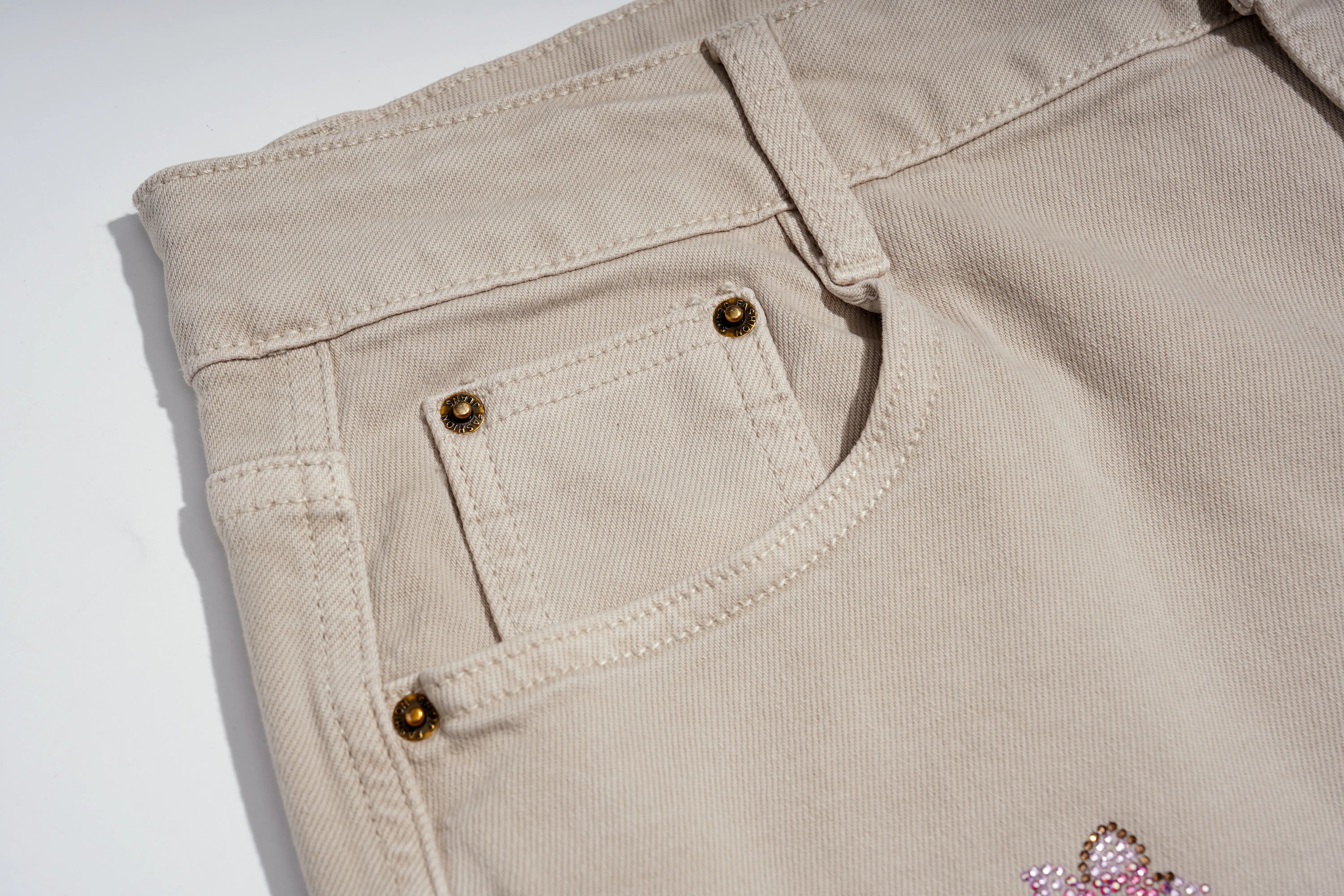 Close-up of the back pocket area of light beige denim jeans, showing brass-colored rivets and a small pink embellished detail on the lower right leg. TCH Rhinestone Cherry Blossom Straight-Leg Jeans