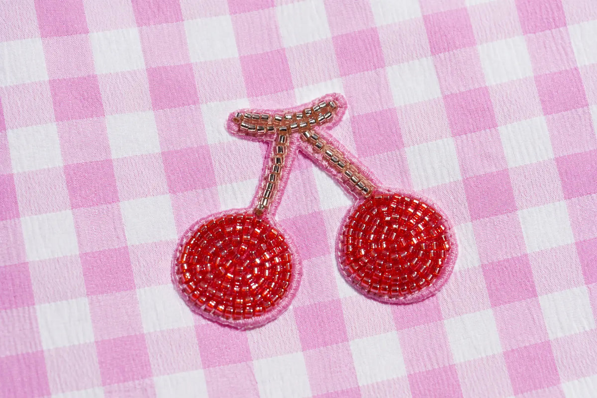 A pair of cherry-shaped beaded earrings with red seed beads, pink borders, and gold-toned stems, displayed on a pink and white checkered fabric background. TCH Rhinestone Cherry Gingham Cargo Wide-Leg Pants