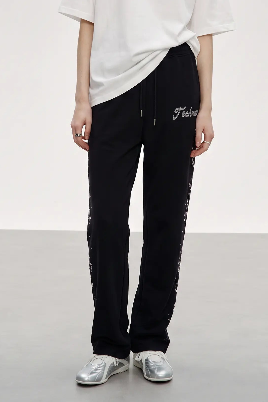 A person wearing black sweatpants with white script embroidery on the thigh and white side graphics, paired with a white t-shirt and silver sneakers, photographed in a studio setting. TCH Rhinestone Constellation Straight-Leg Sweatpants