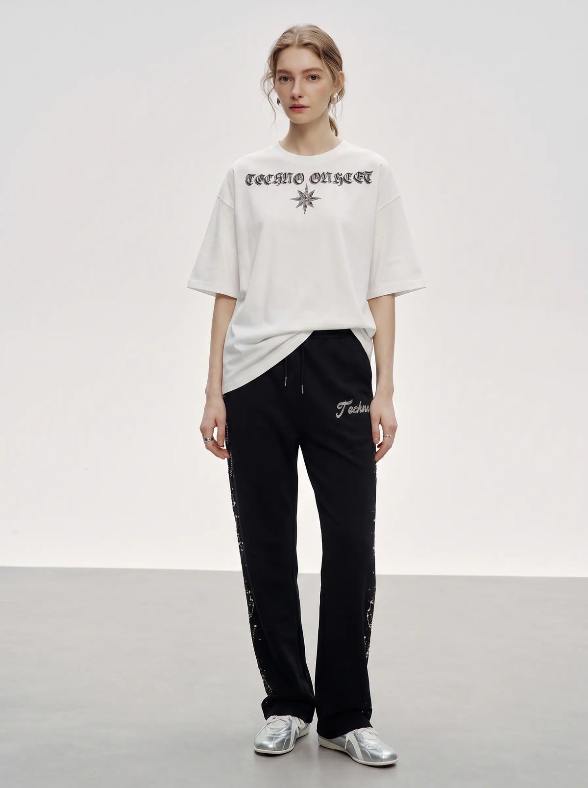 A model stands in a studio wearing a white oversized T-shirt with black gothic text and a star graphic, paired with black pants featuring white script and side print details, and silver sneakers. TCH Rhinestone Constellation Straight-Leg Sweatpants