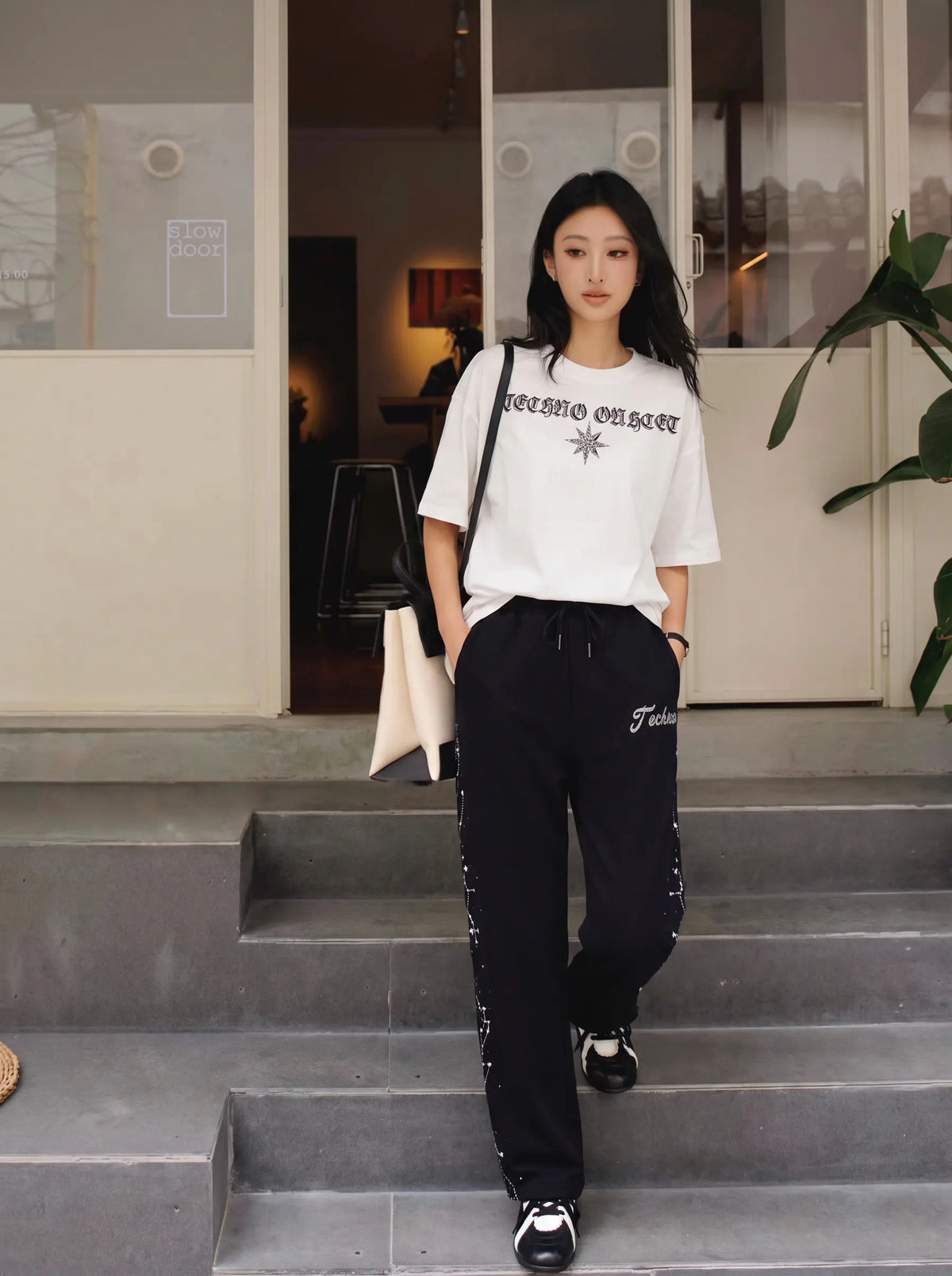 A woman stands on outdoor concrete steps wearing a white graphic t-shirt and black sweatpants with white lettering, carrying a white tote bag. TCH Rhinestone Constellation Straight-Leg Sweatpants