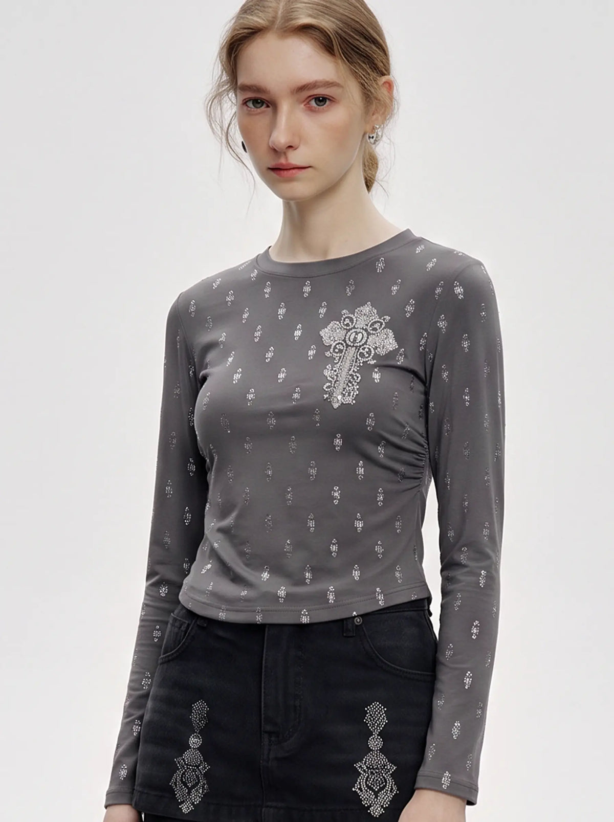 A woman modeling a fitted long-sleeve gray top with a white embroidered cross motif on the chest and small scattered white embroidered details, paired with black skirt or shorts featuring white embroidery. TCH Rhinestone Cross Graphic Fitted Long Sleeve Top