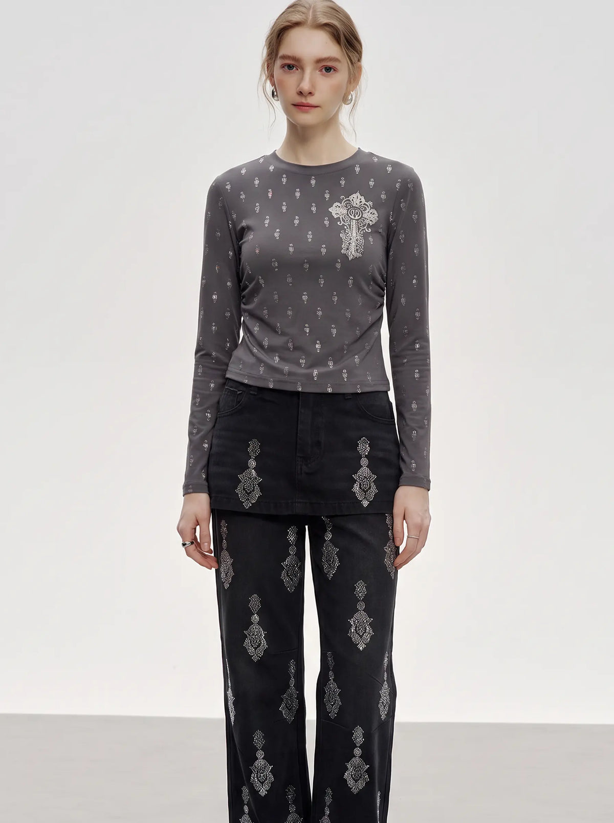 A model stands against a light gray studio background wearing a dark gray long-sleeve top with a small white embroidered cross motif on the left chest, paired with black wide-leg pants featuring white embroidered details. TCH Rhinestone Cross Graphic Fitted Long Sleeve Top