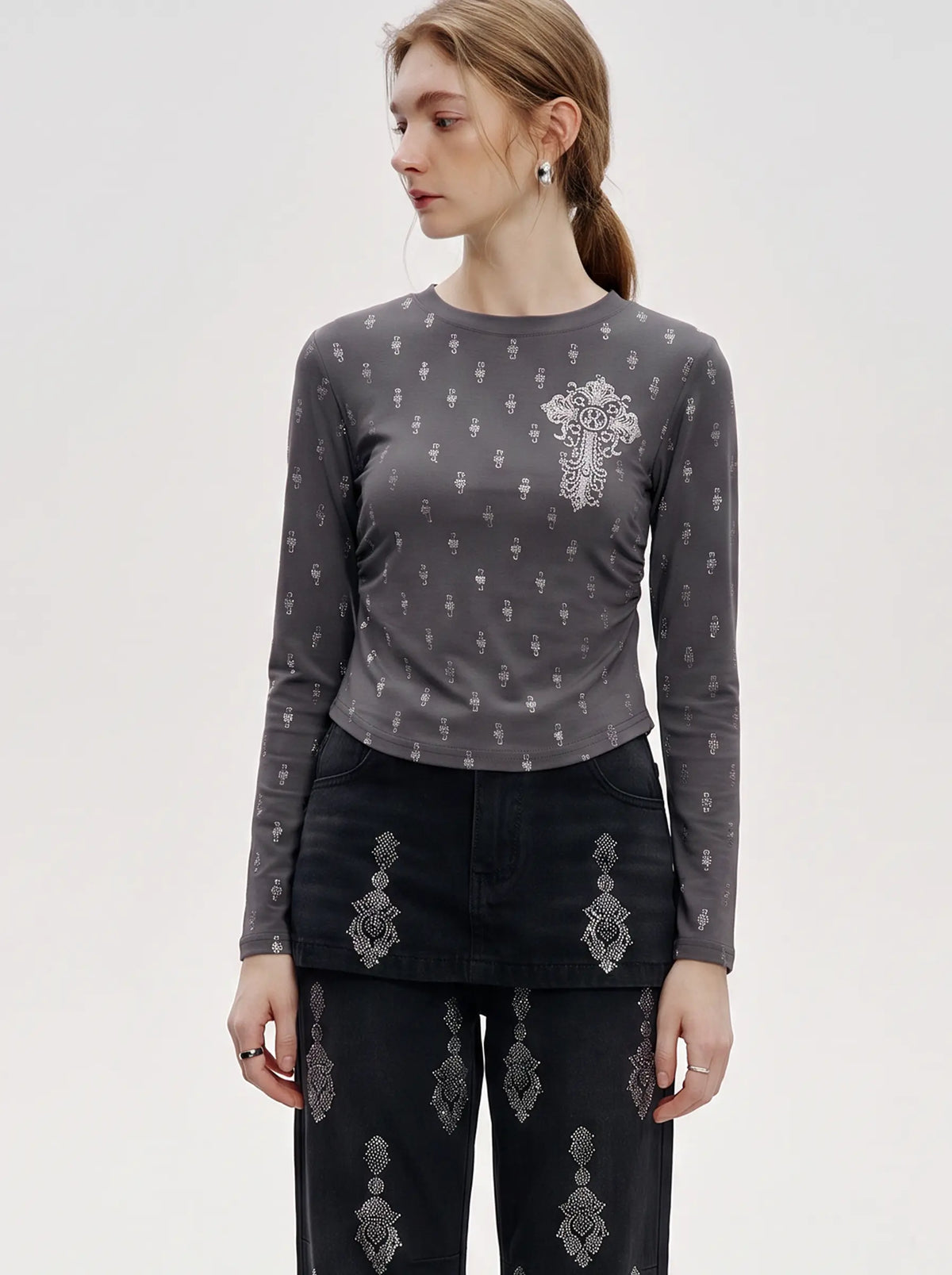 A woman modeling a dark gray long-sleeve top with a small white cross and vertical cross pattern, paired with black pants featuring white cross motifs, against a plain light gray background. TCH Rhinestone Cross Graphic Fitted Long Sleeve Top